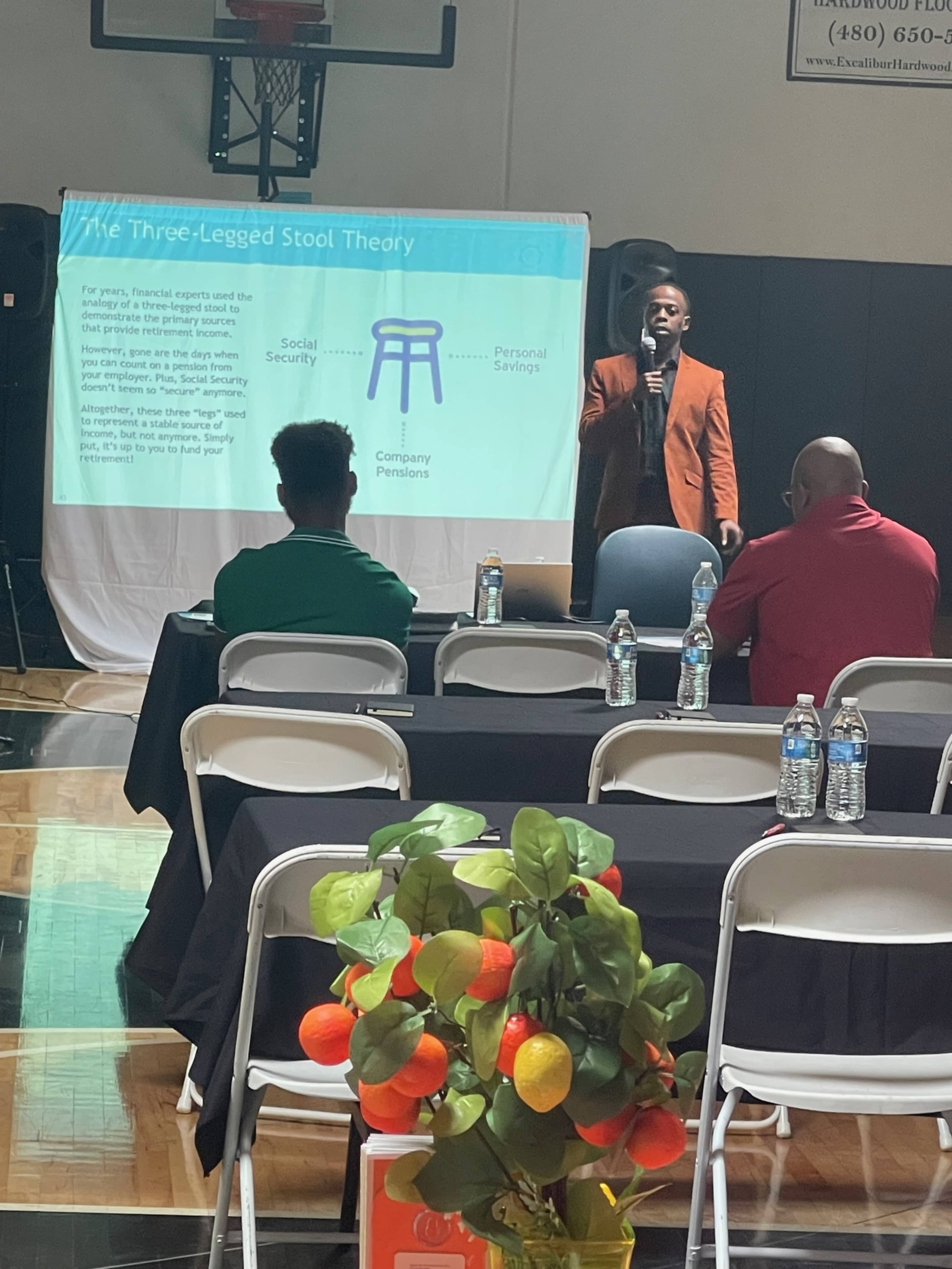 A speaker presents the "Three-Legged Stool Theory" on a projector screen in a gymnasium setting while an audience watches from their seats.