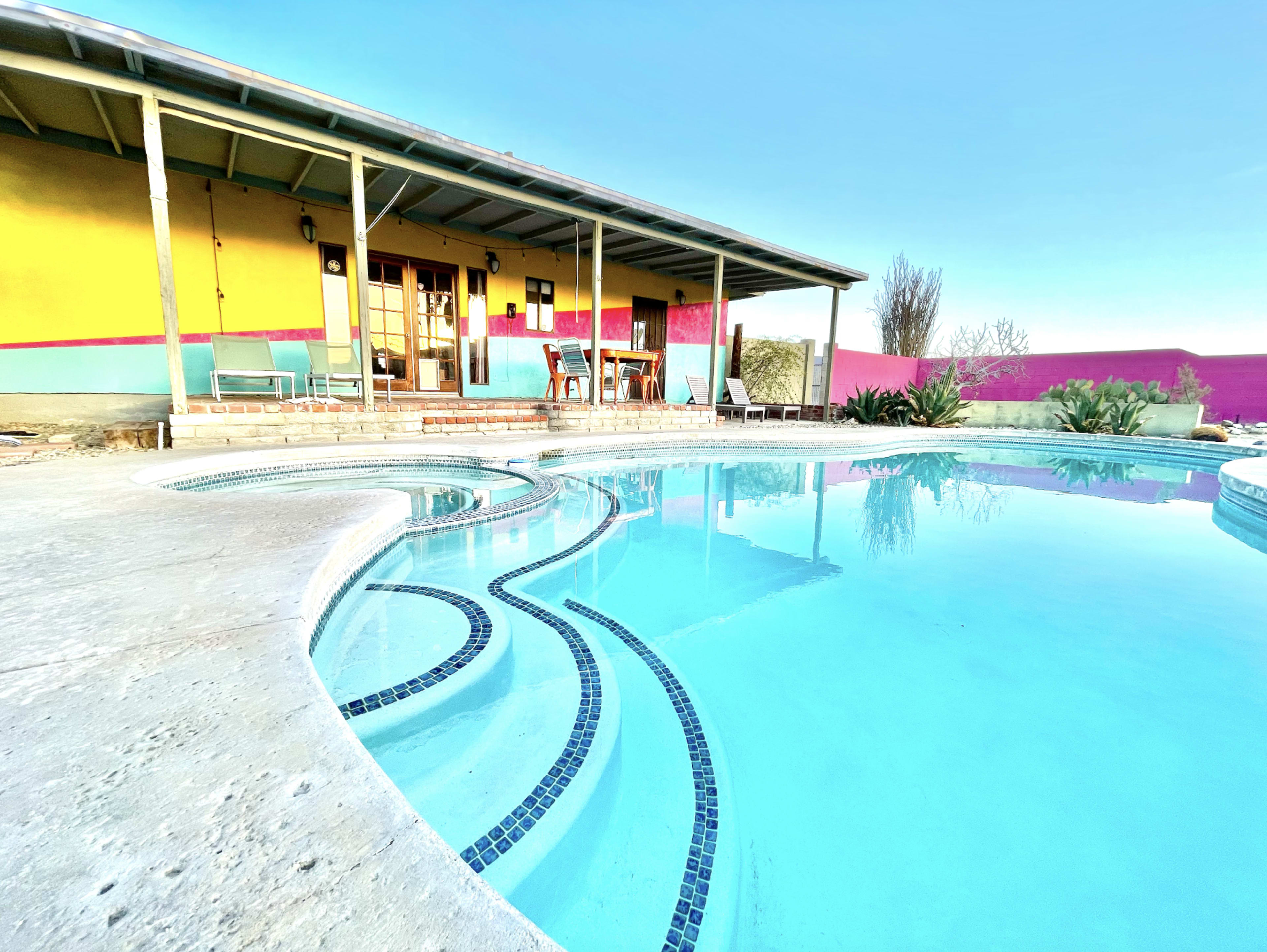 A swimming pool with a patterned edge is situated in front of a colorful house with a covered porch.