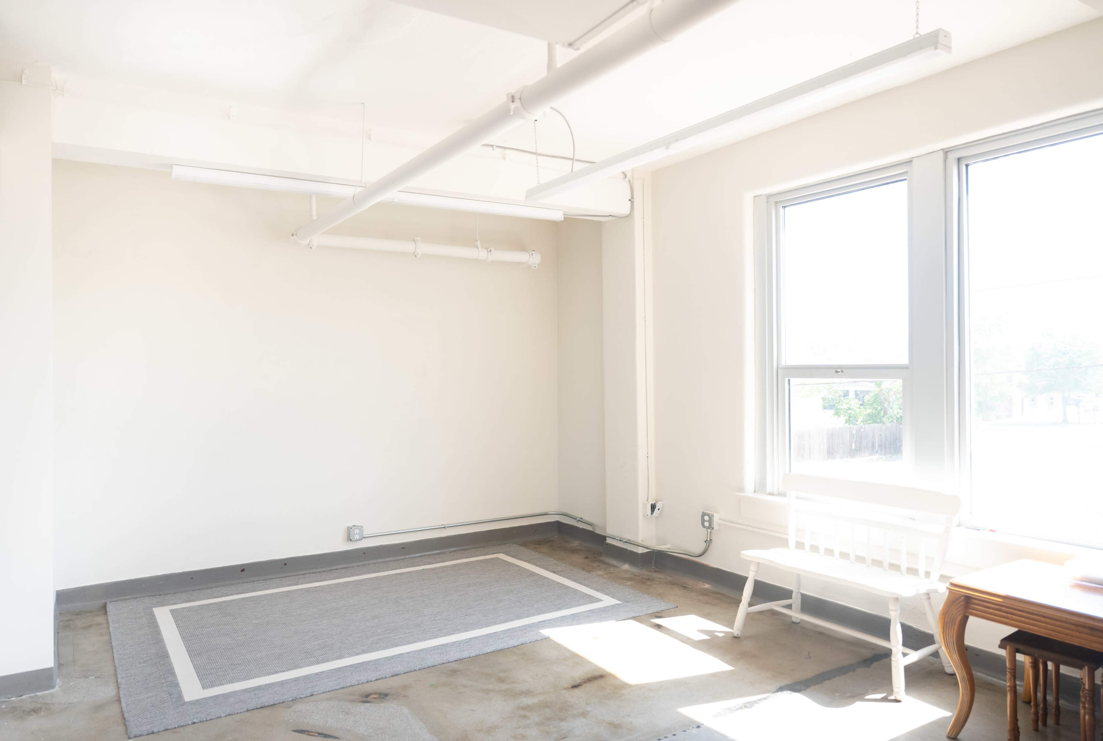 The image shows a minimalist room with a concrete floor, a small white bench, a large window allowing natural light, and a gray area rug on the floor.