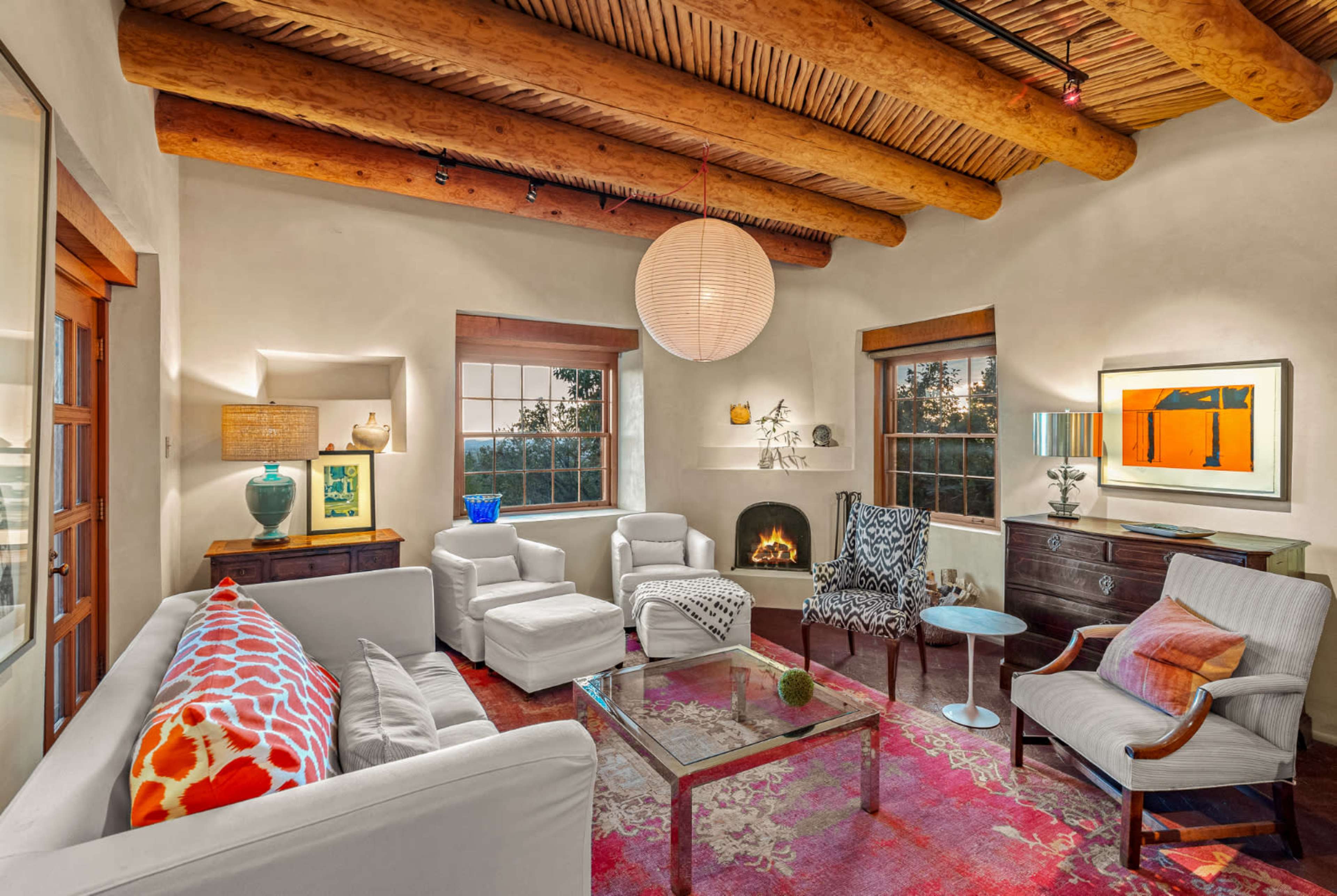 A cozy living room features a mix of white furniture and colorful accents, with wooden beams on the ceiling and large windows providing natural light.