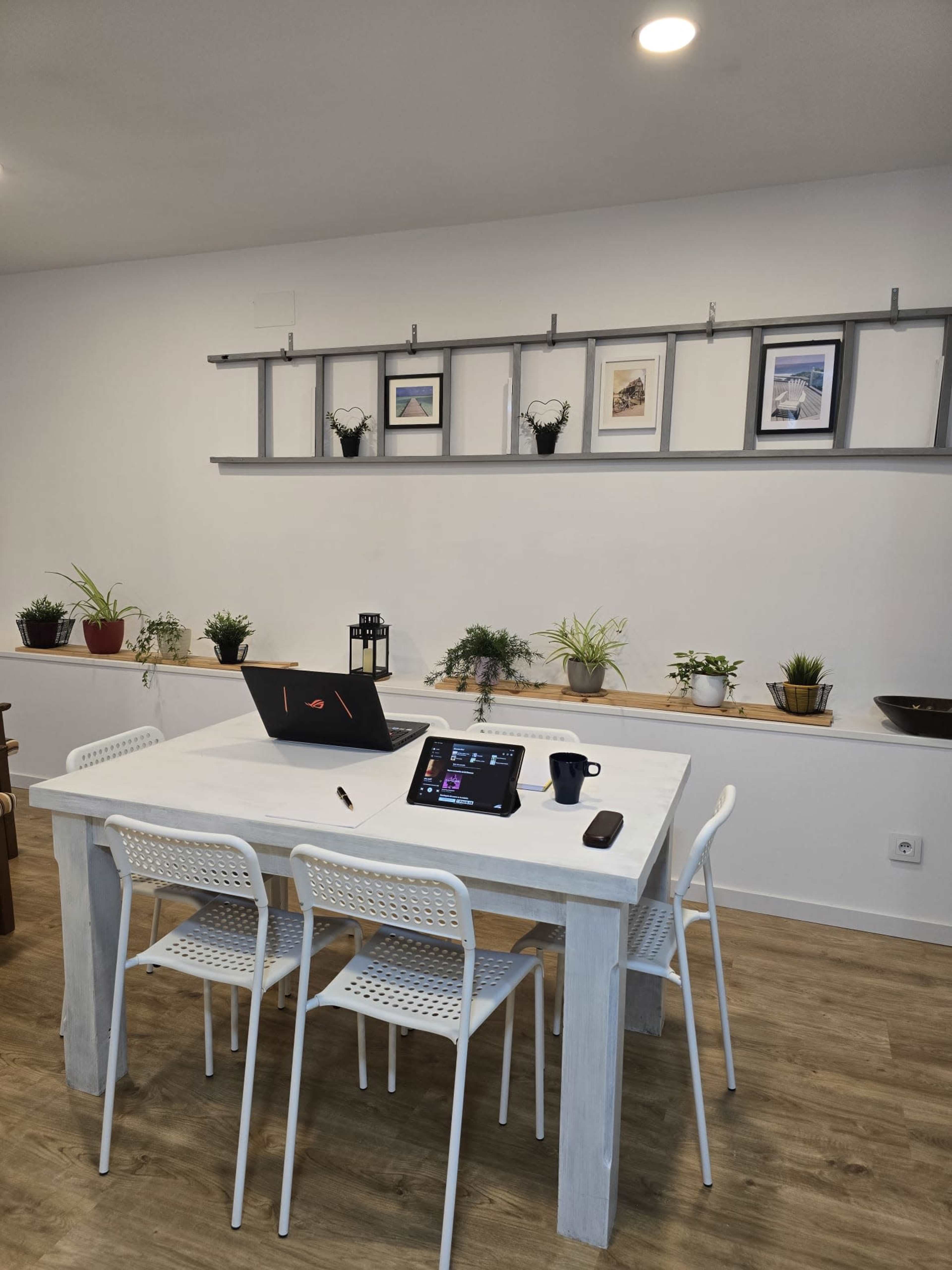 A modern dining area features a white table with four chairs, a laptop, a tablet on the table, and potted plants lining the wall-mounted shelf.