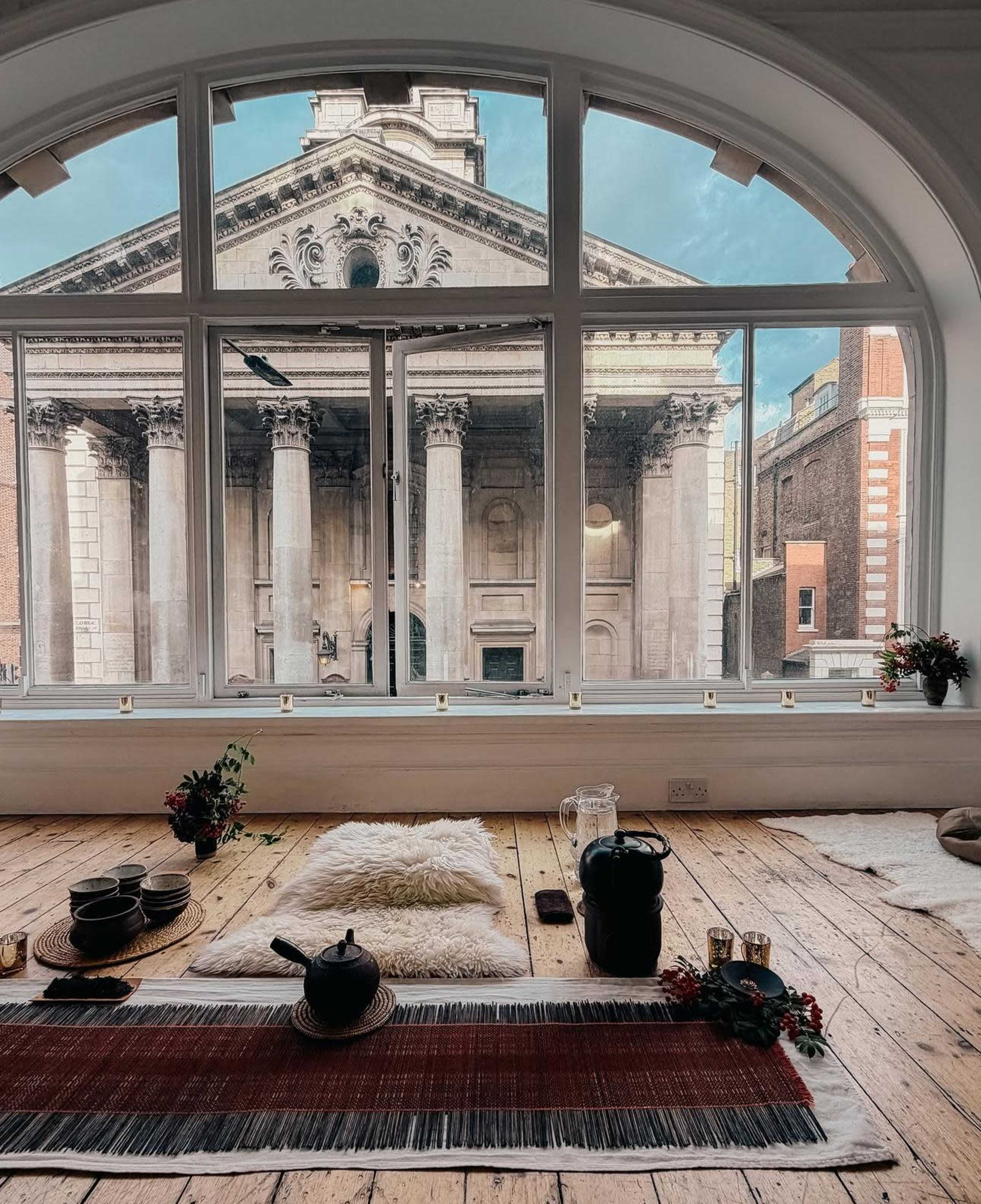 The scene features a window view of a grand building with columns, while a cozy, minimalist interior with cushions and tea accessories is foregrounded.