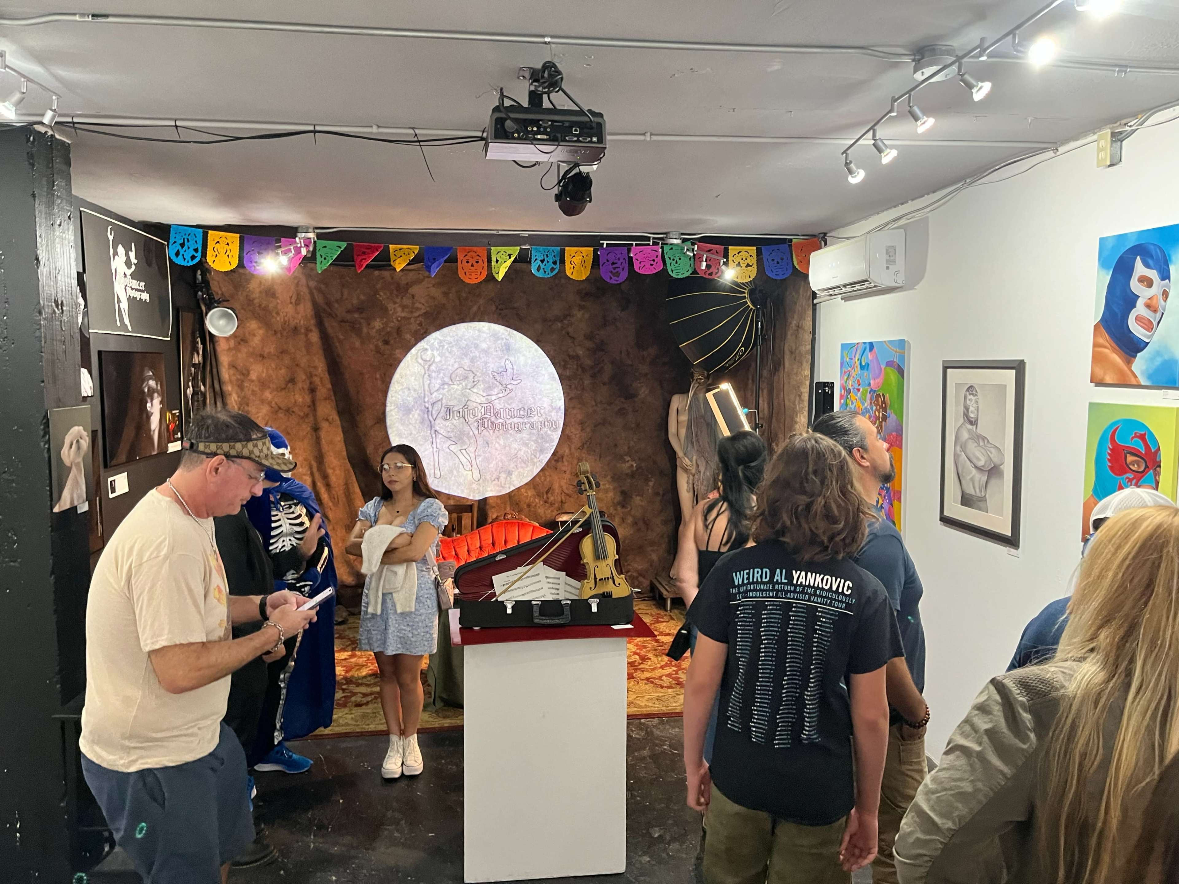 The image shows a vibrant art gallery featuring a diverse group of people admiring various artworks, including paintings and sculptures, with colorful decorations and a prominent circular backdrop.