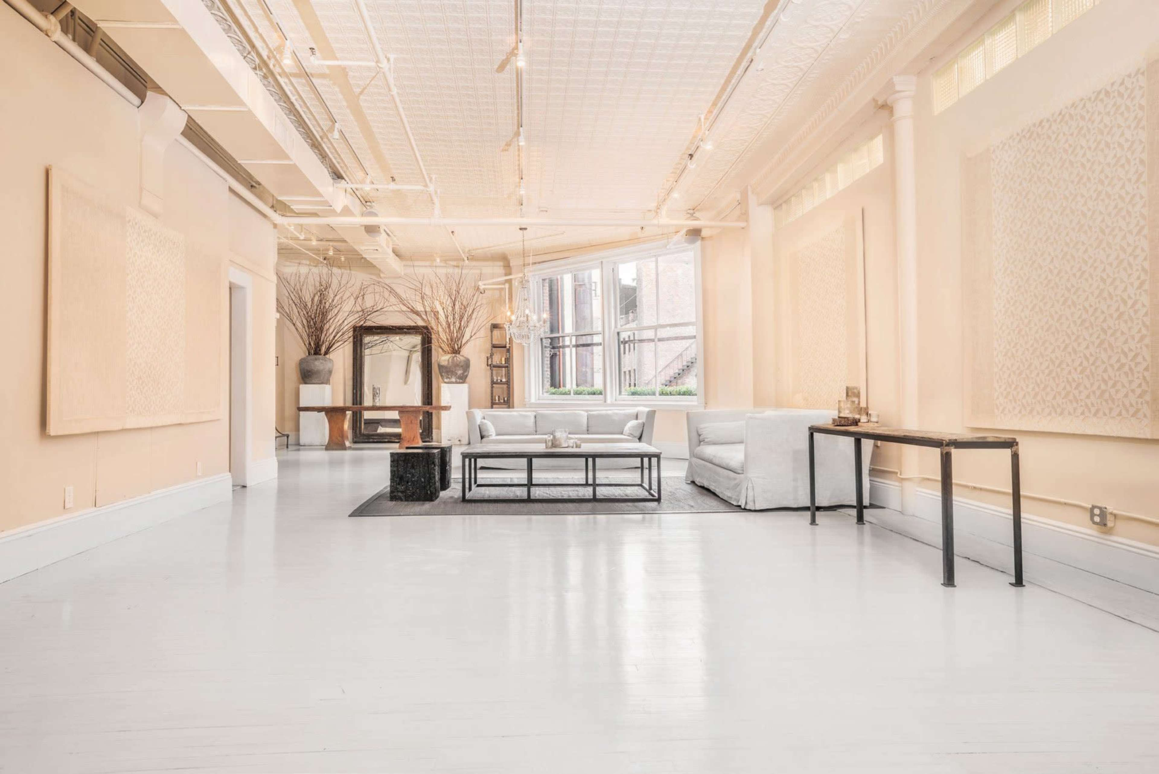 The image shows a spacious, well-lit room with light-colored walls, a large window, minimal furniture including a couch and a coffee table, and decorative elements like a mirror and plants.