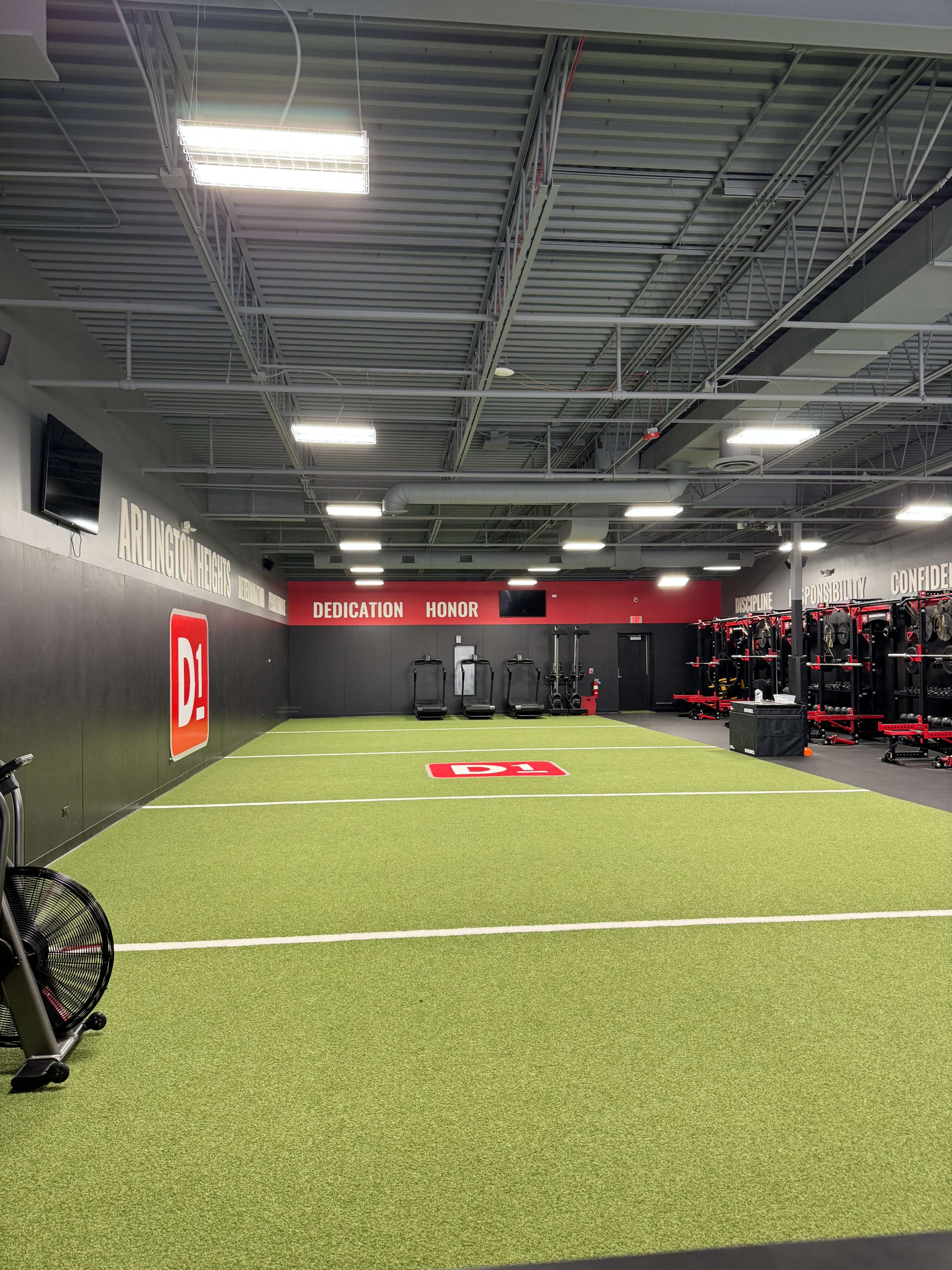 The image shows a well-lit training facility featuring artificial turf, exercise equipment, and motivational signage on the walls.
