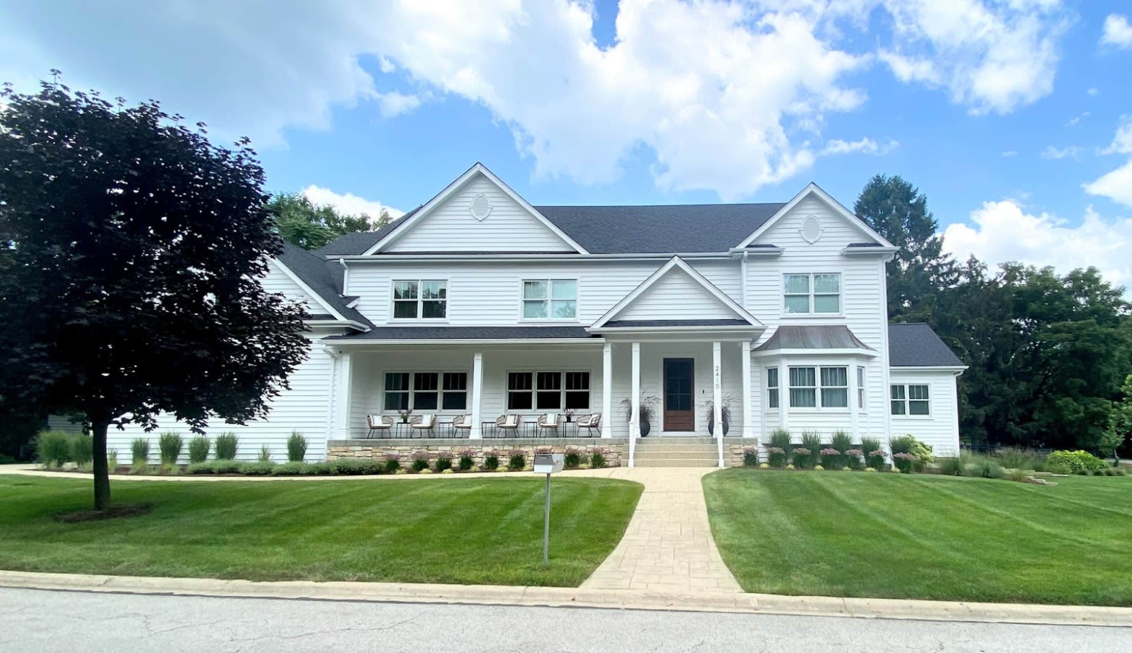 The image shows a large, white two-story house with a front porch, well-manicured lawn, and a sidewalk in a suburban neighborhood.