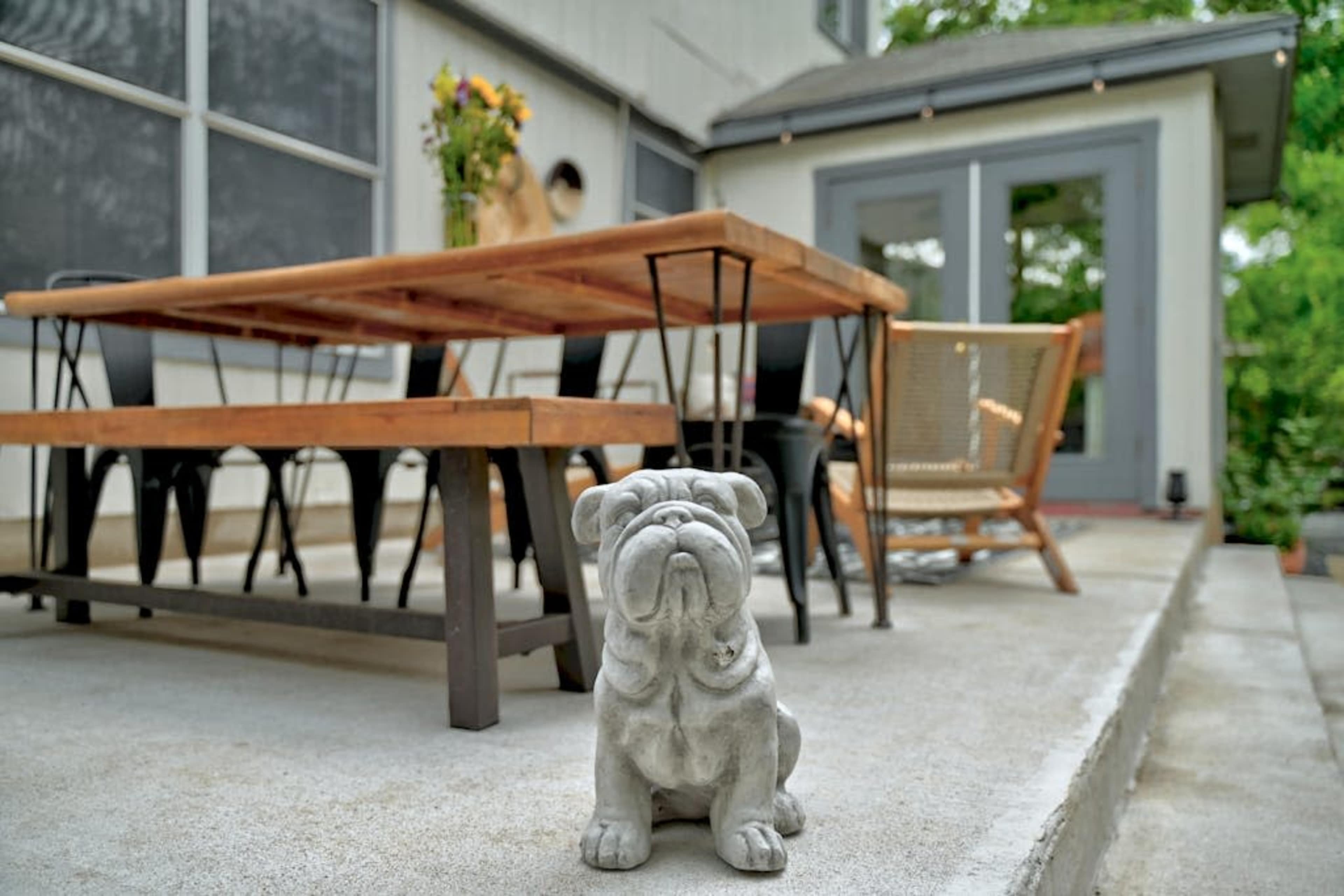 A concrete bulldog statue sits on a patio in front of a wooden dining table and chairs next to a house.