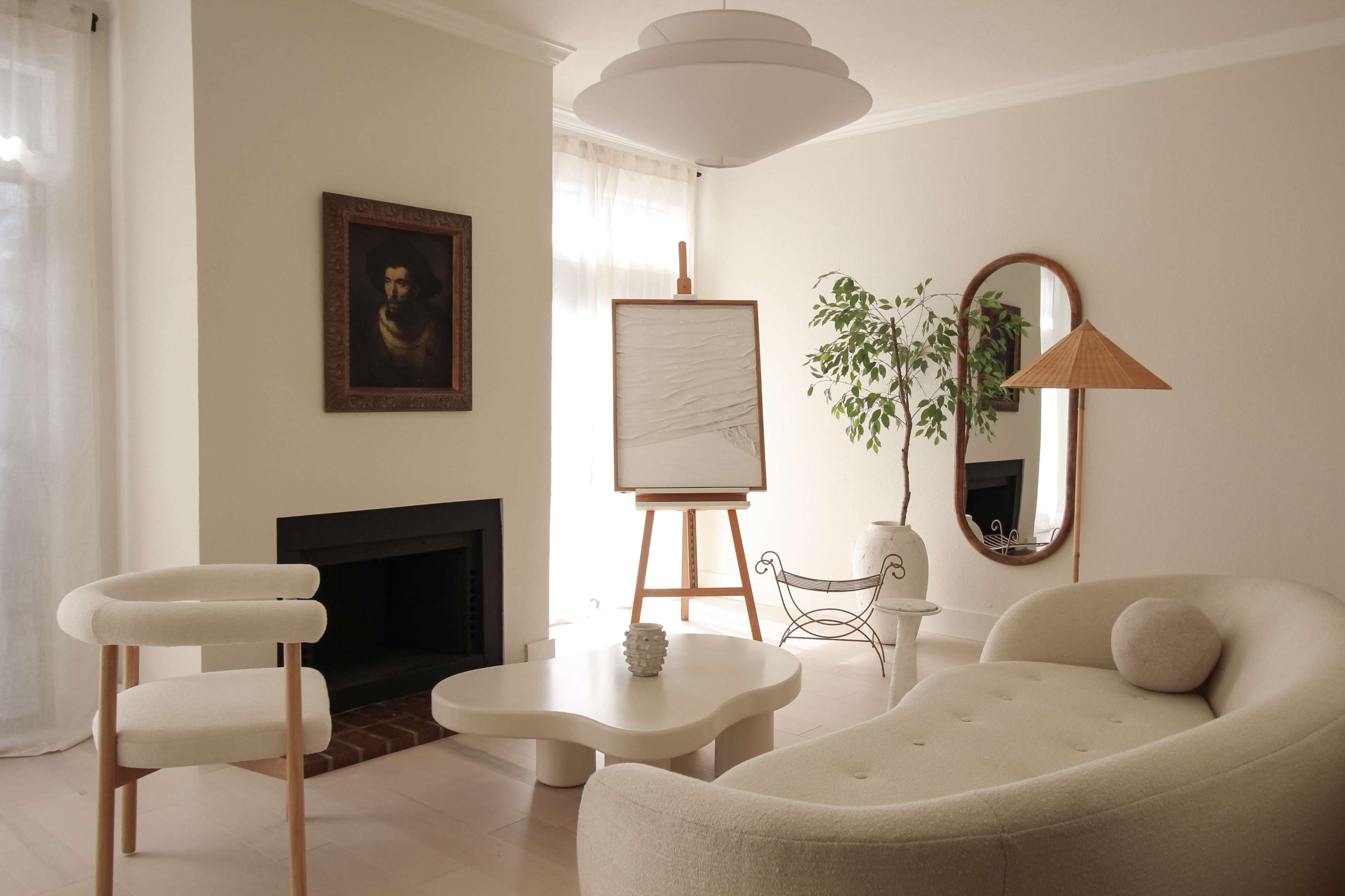 The image depicts a modern living room featuring a light-colored sofa, a minimalistic coffee table, an easel with a blank canvas, and a large indoor plant.