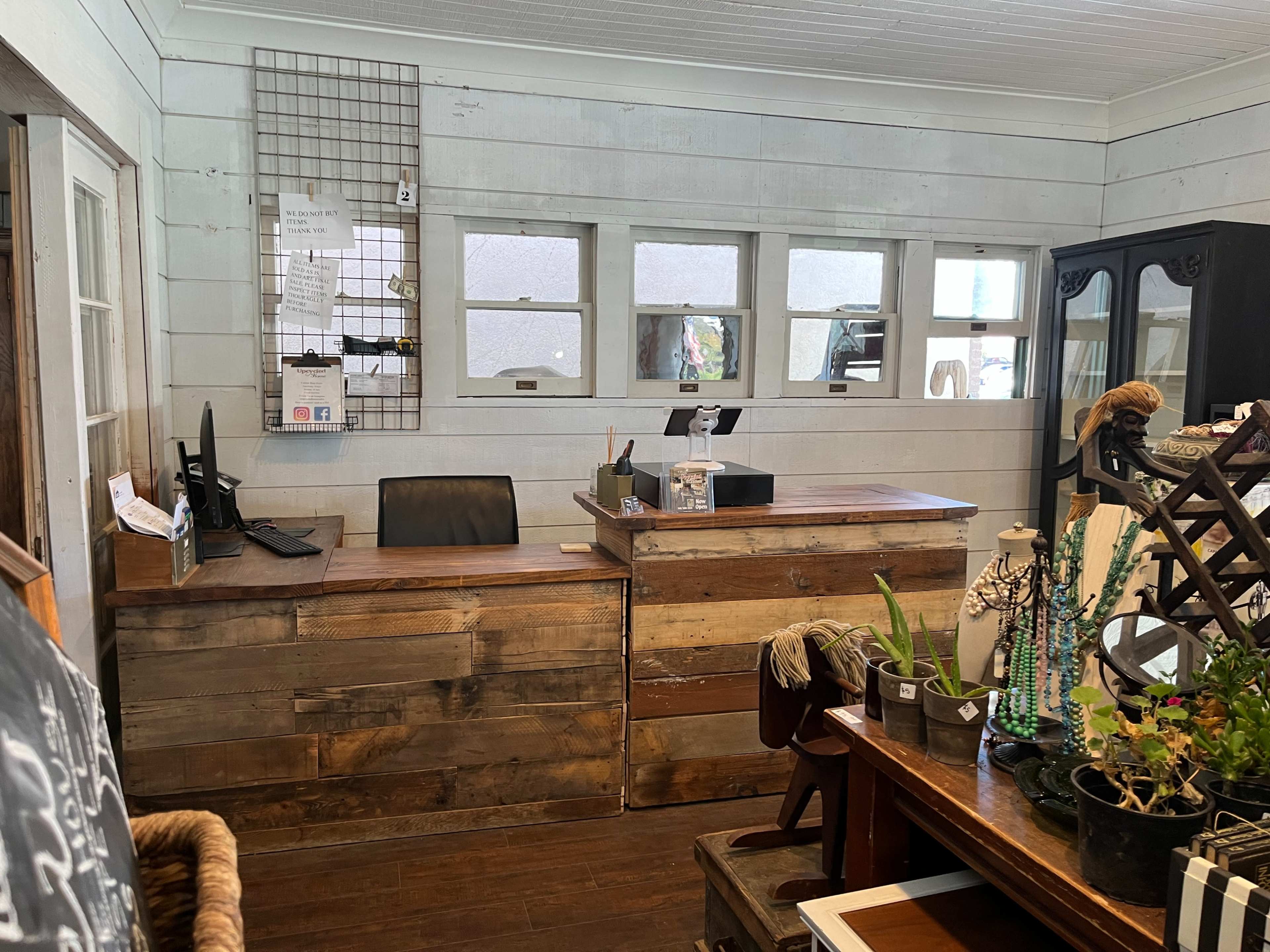 A wooden reception area with a desk, computer, and decorative plants against a backdrop of whitewashed walls and several small windows.