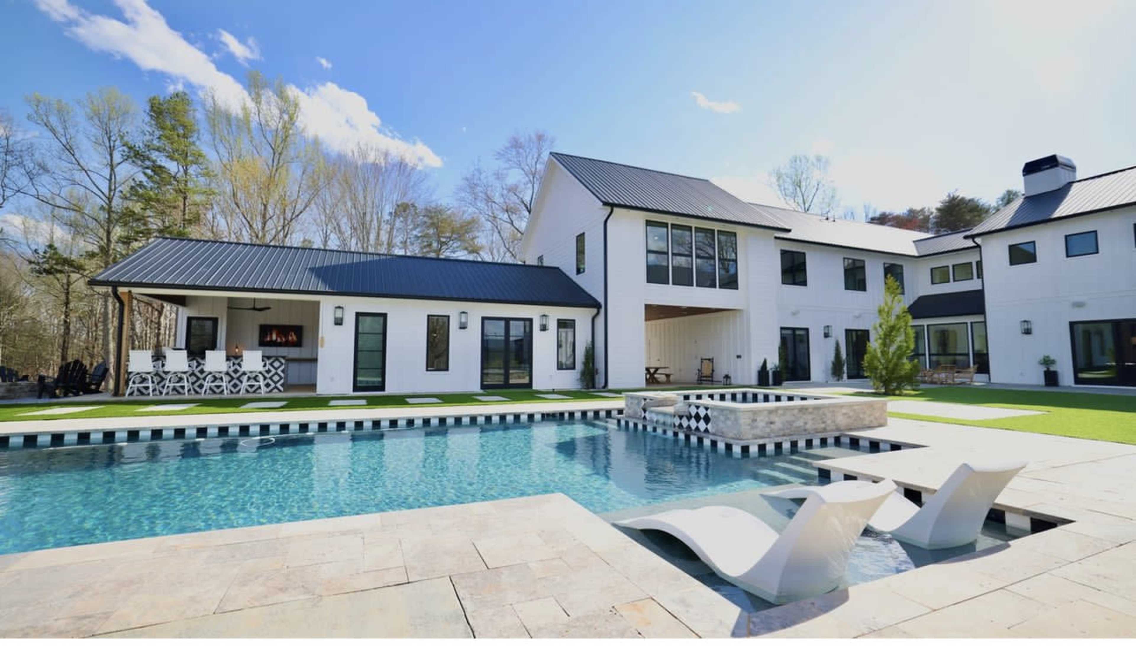 A modern two-story house with large windows and a black metal roof is situated beside a swimming pool featuring a spa and lounge chairs.