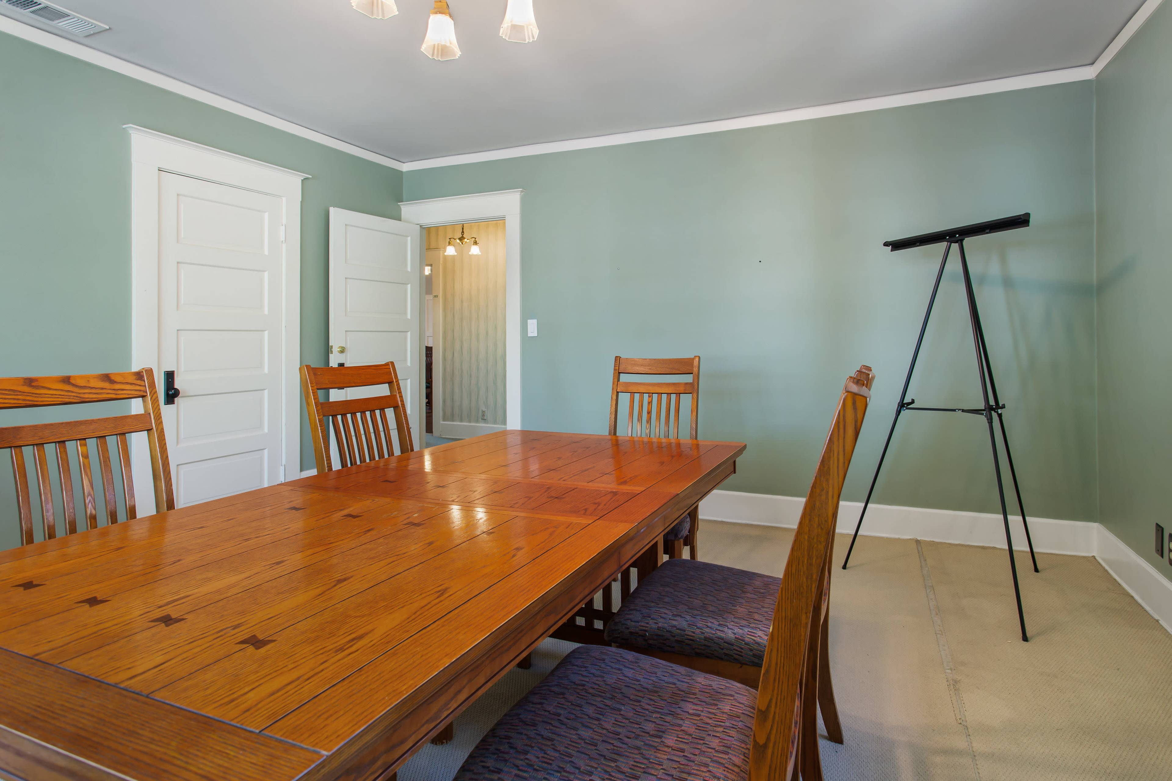 A wooden dining table with chairs is positioned in a light green room, featuring a doorway and an easel against the wall.