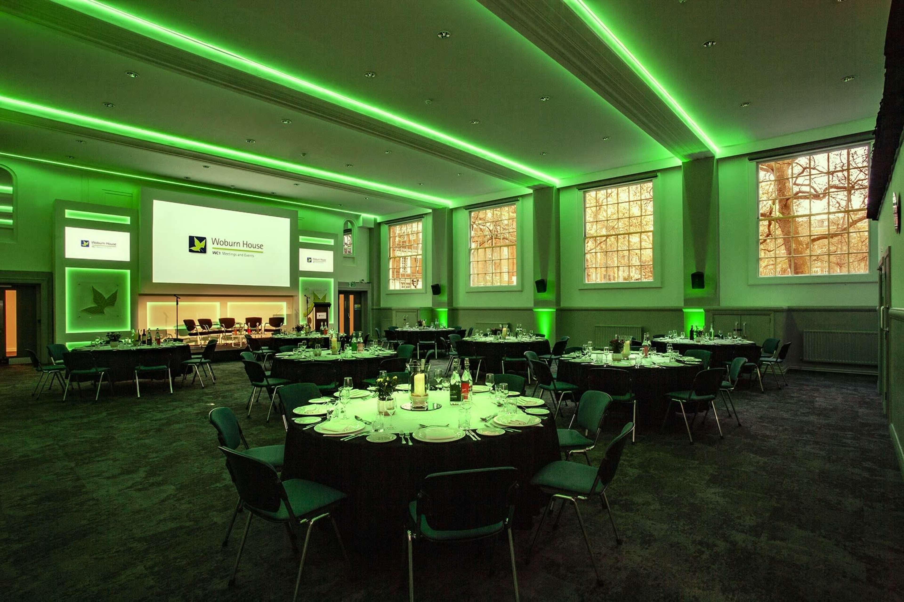 A banquet hall featuring round tables set for dining, illuminated with green lighting and equipped with a large screen for presentations.