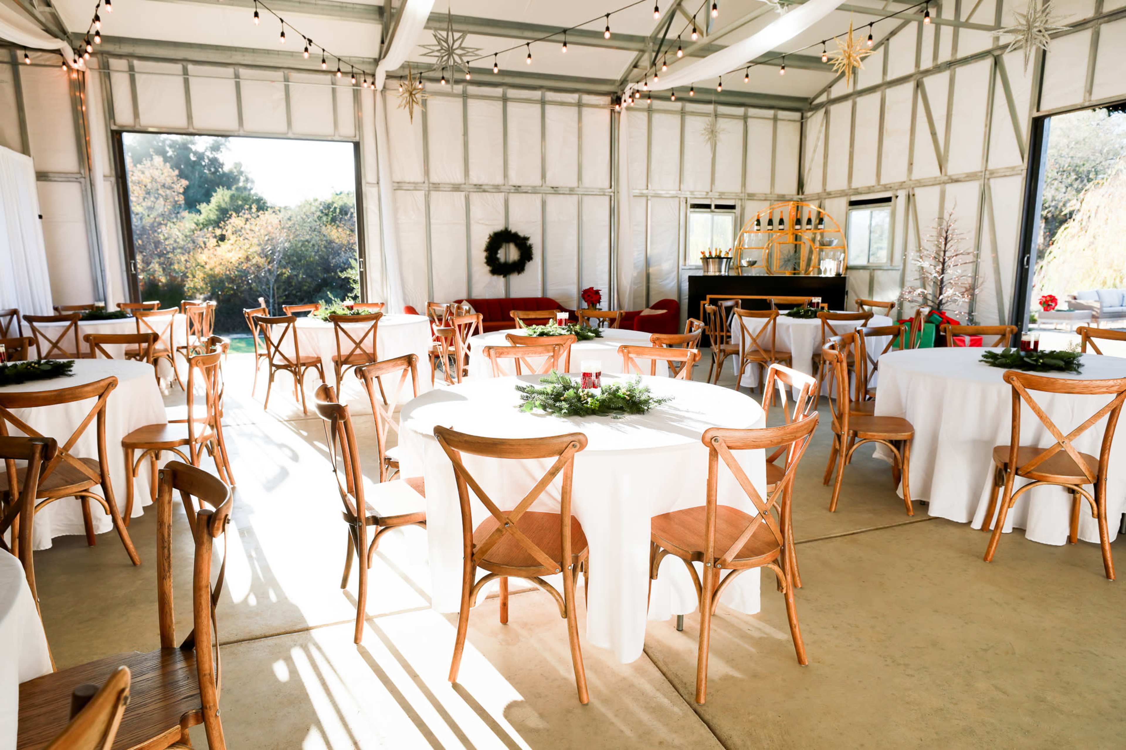 A spacious event venue with round tables adorned with white tablecloths and greenery, featuring wooden chairs and festive decorations.