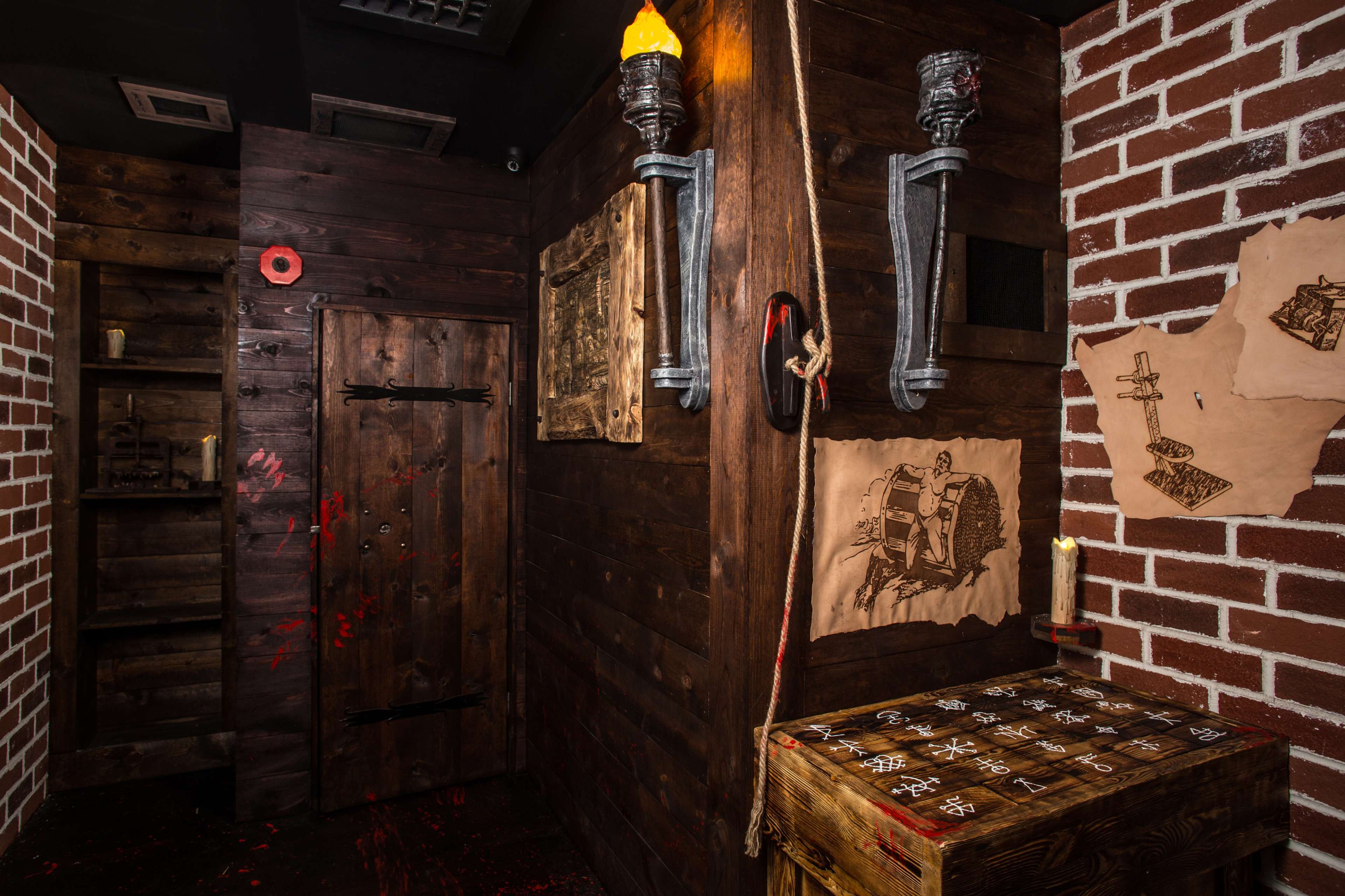 A dimly lit room with wooden walls, a door, and various decorations including torches, old maps, and a wooden table with markings.