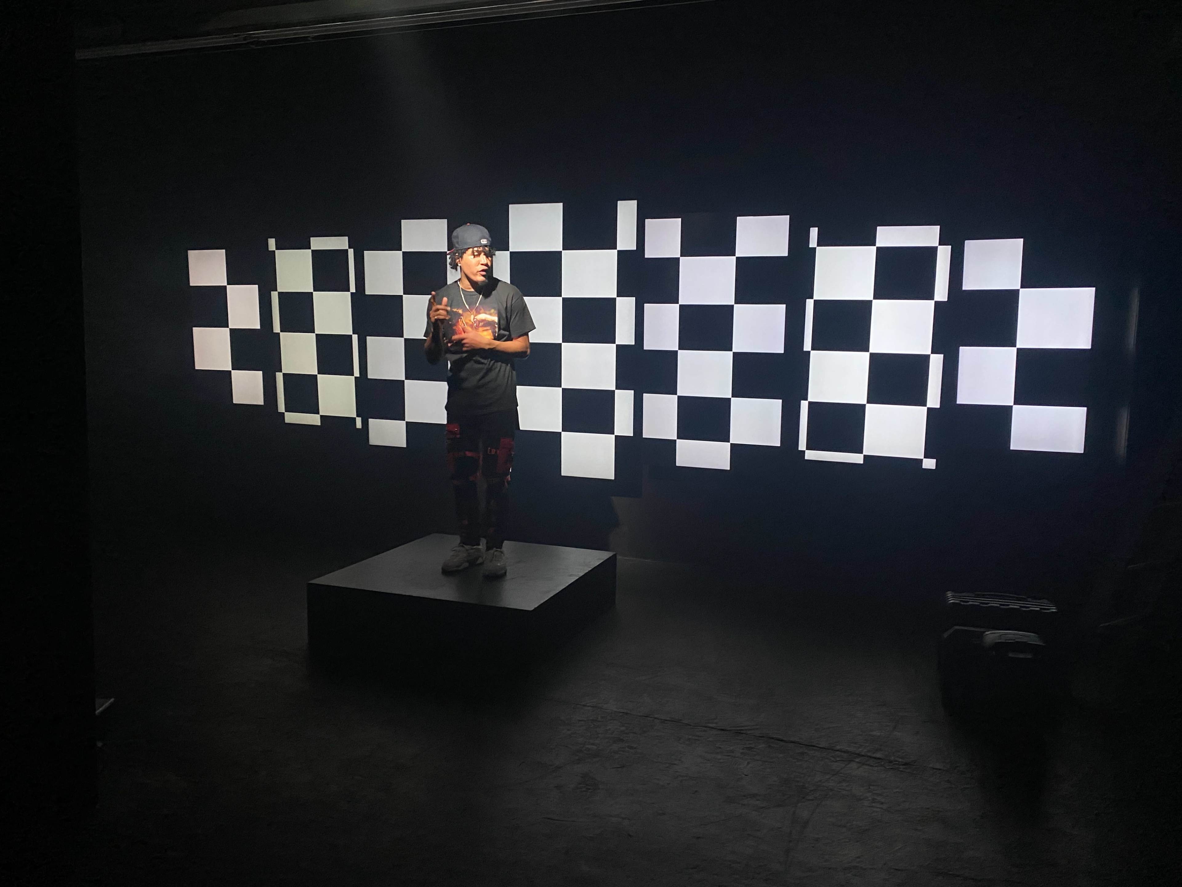 A person stands on a platform in a dark room, surrounded by a backdrop of alternating light and dark squares.