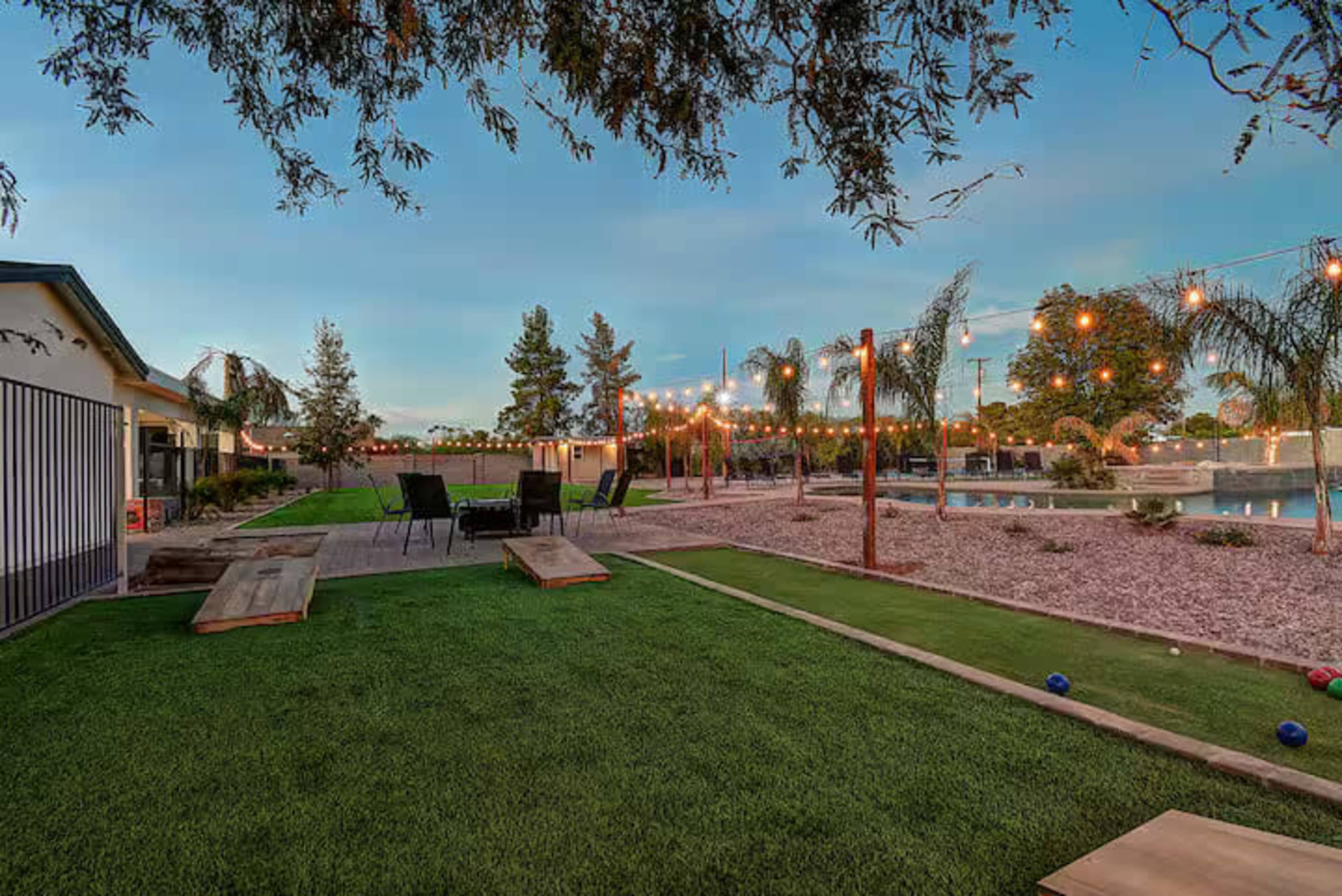 The image shows a landscaped outdoor area with string lights, seating arrangements, palm trees, and a swimming pool in the background.