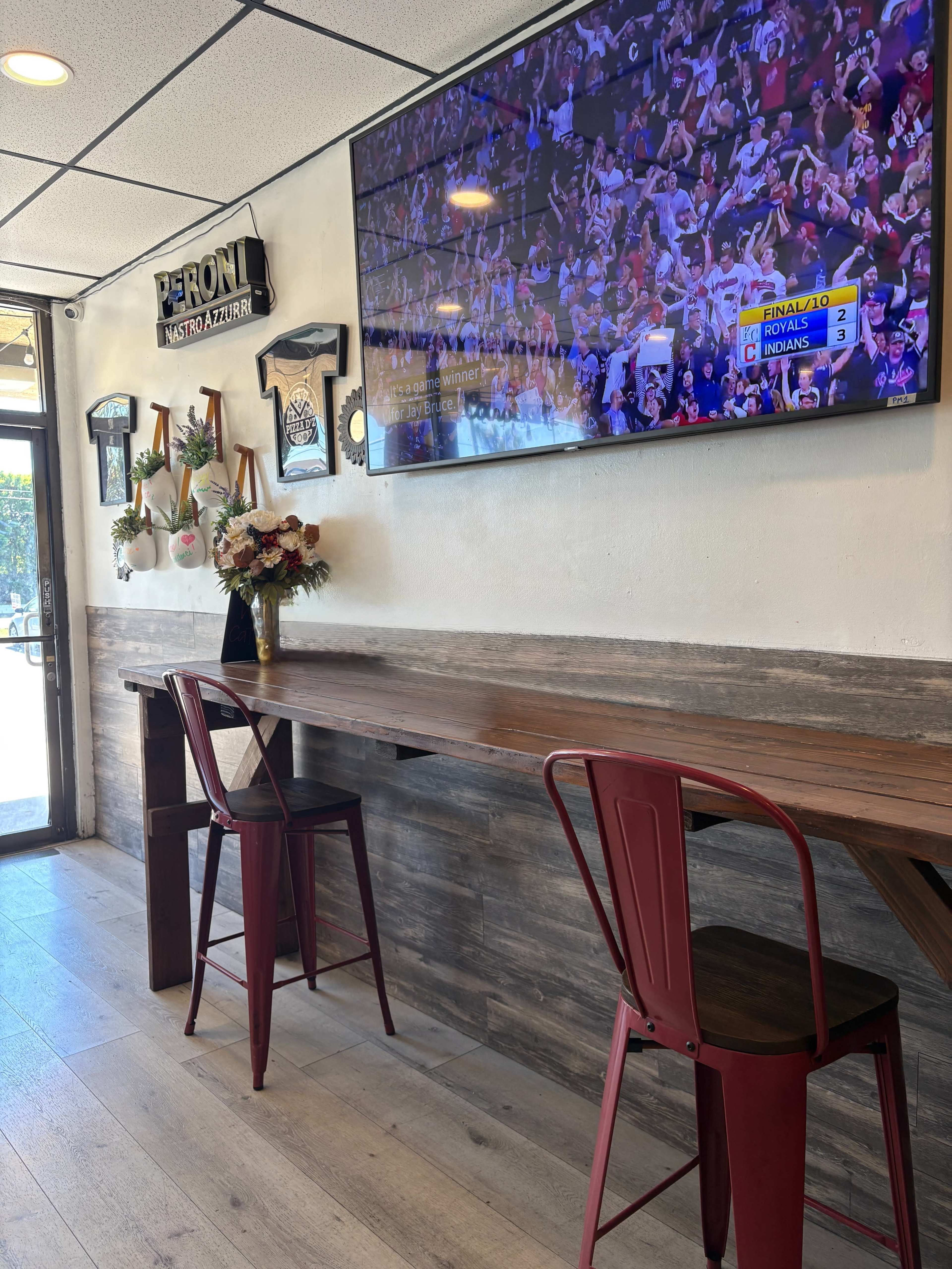 The image shows a restaurant interior with a long wooden table and two red metal chairs, featuring a large television displaying a sports event in the background.