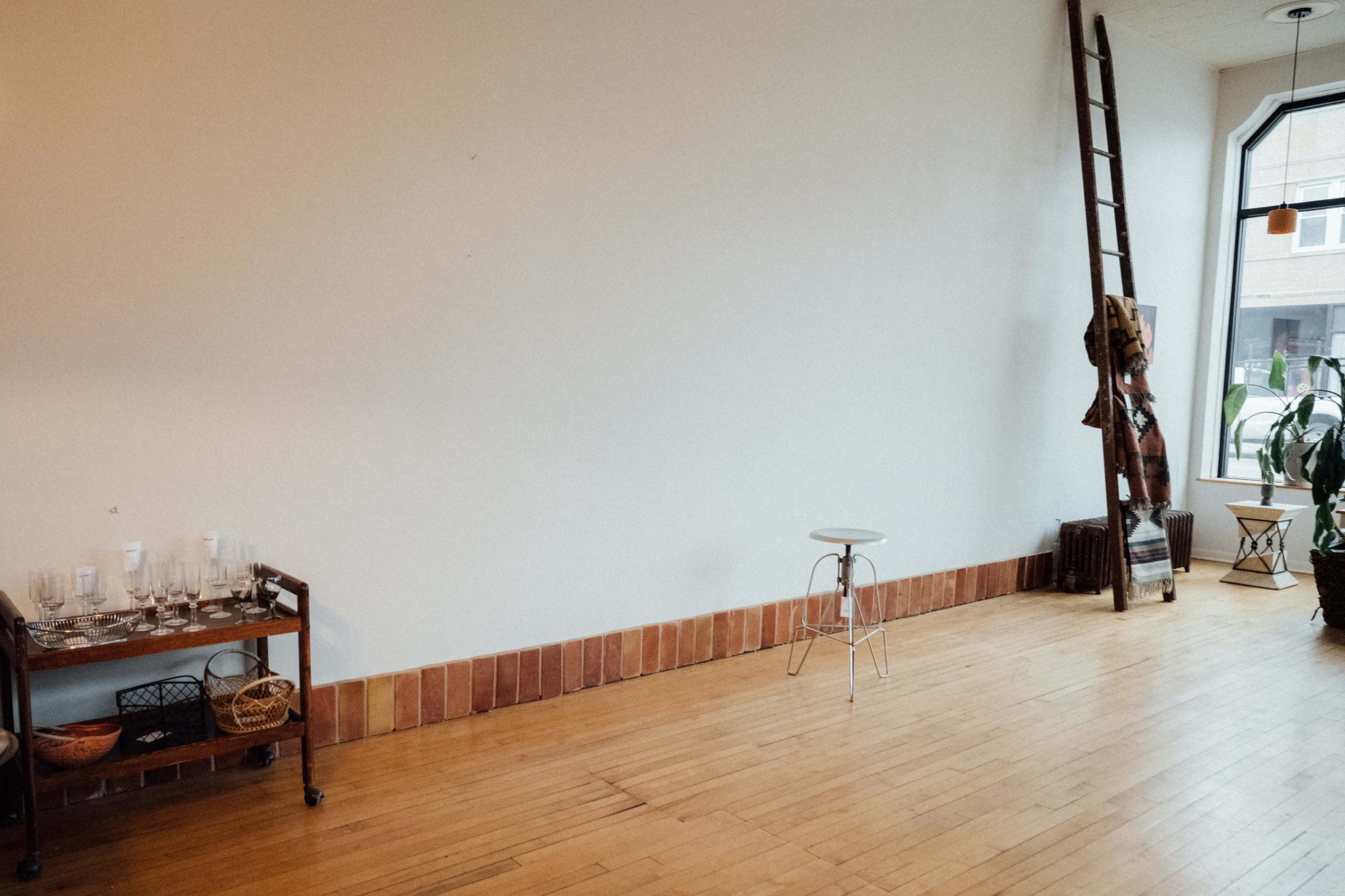 A spacious room features wooden flooring, a blank wall, a tall ladder, a small stool, and a cart with glassware and baskets.