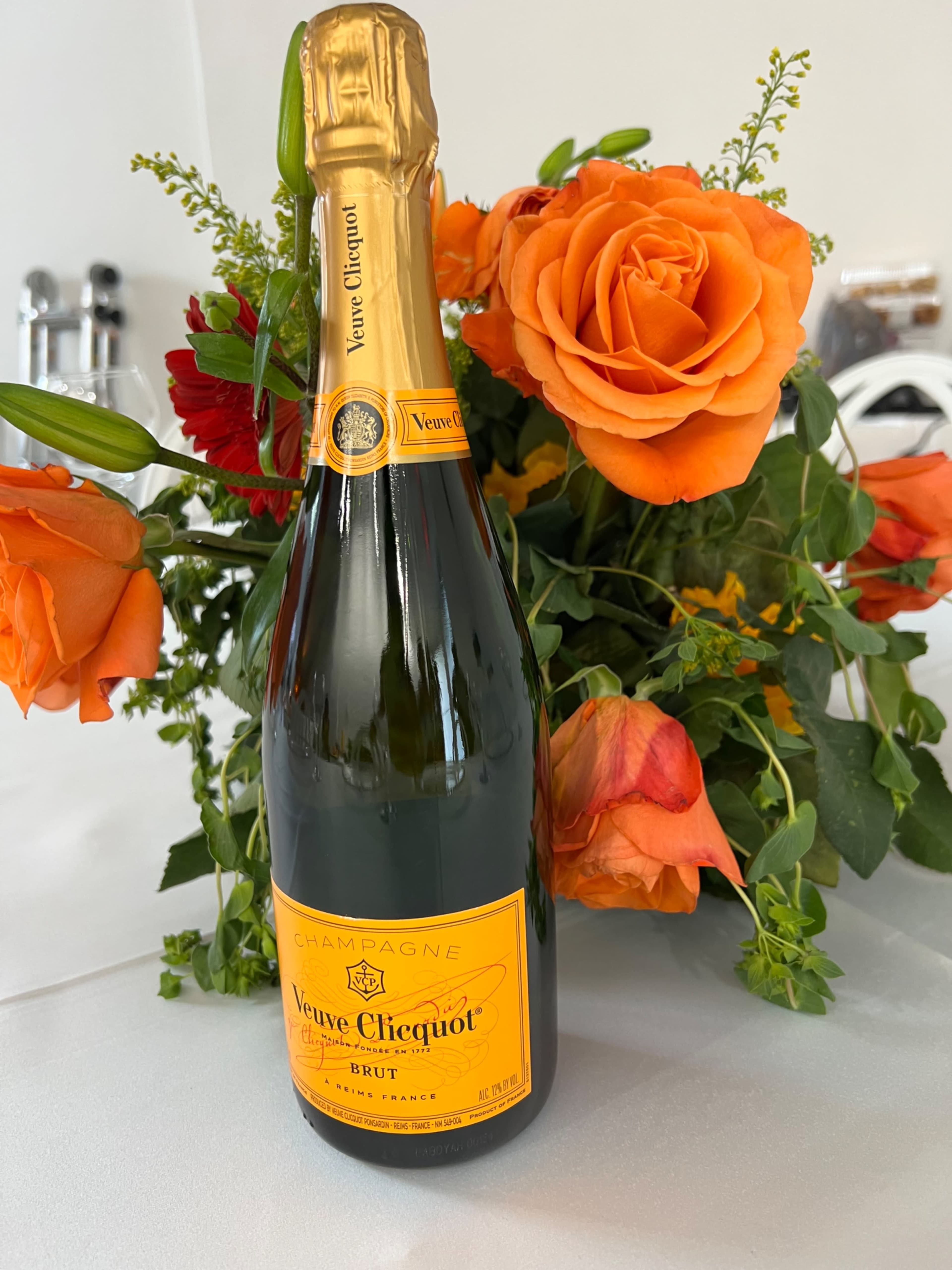 A bottle of Veuve Clicquot champagne is placed in front of a bouquet of orange roses and green foliage.