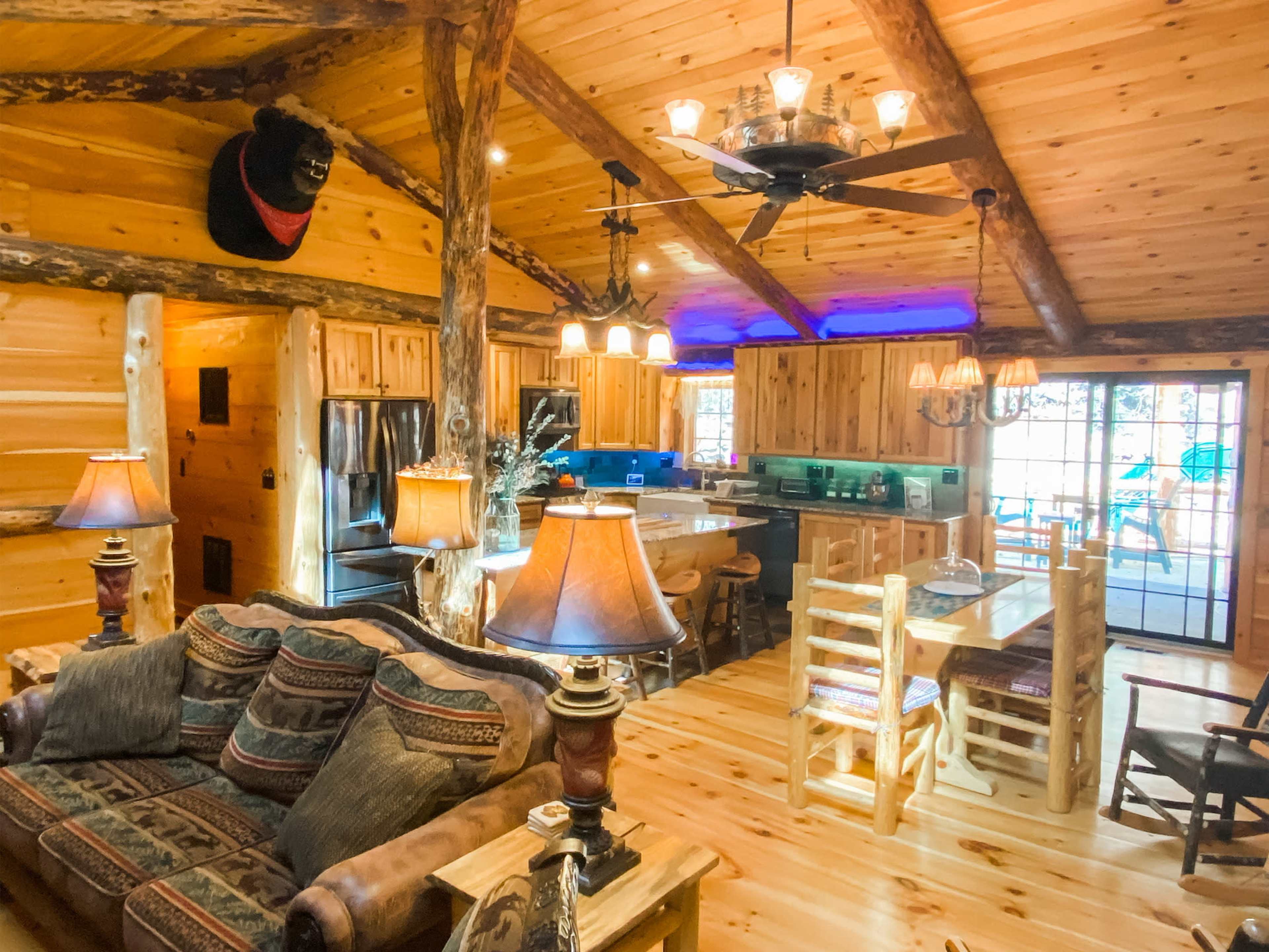 The image shows a rustic wooden cabin interior with a living area featuring a couch, a dining table, and a kitchen area in the background.