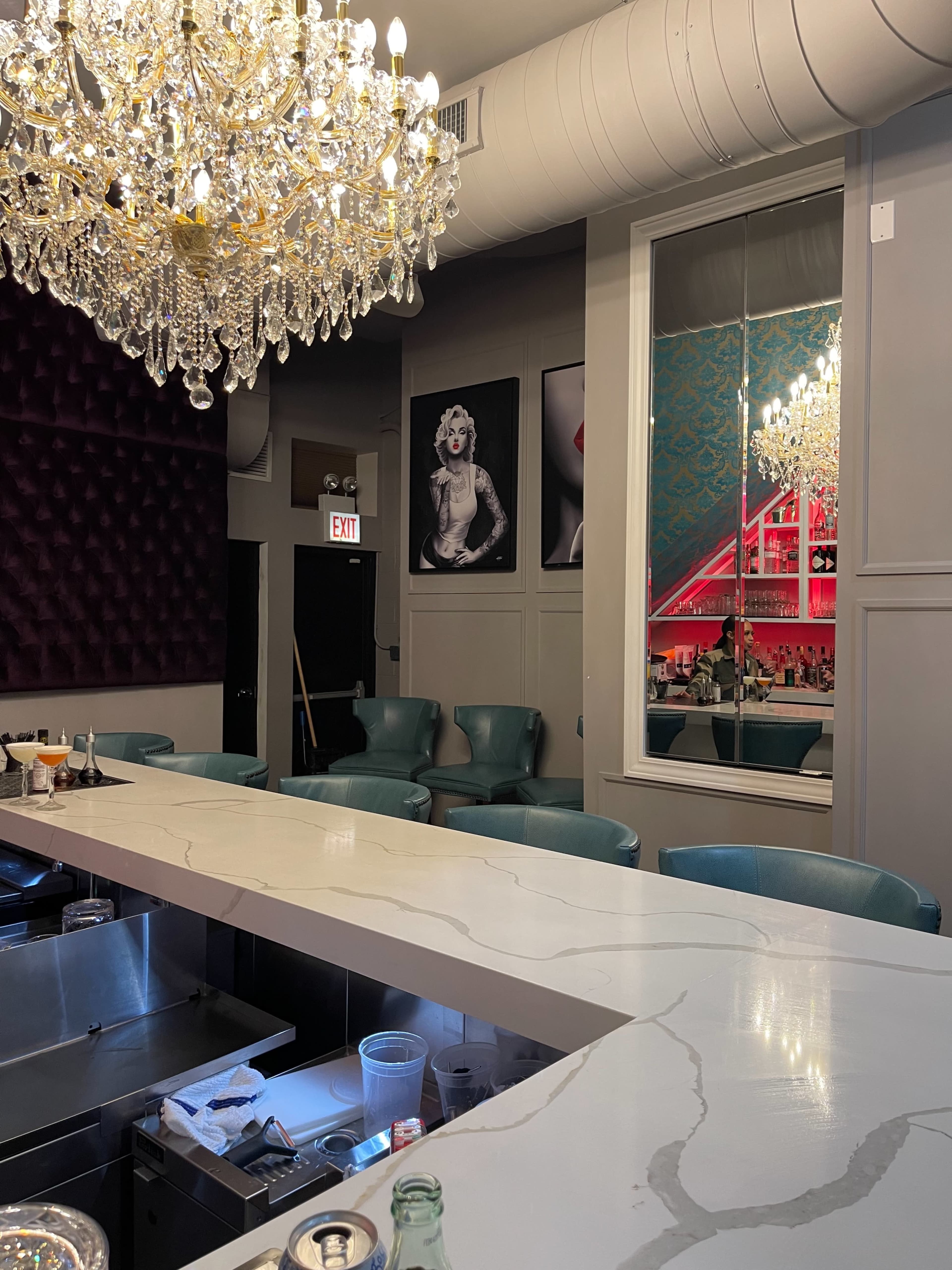 A modern bar with a marble countertop, turquoise chairs, a large chandelier, and a window reflecting artistic wall decor and a well-lit interior.