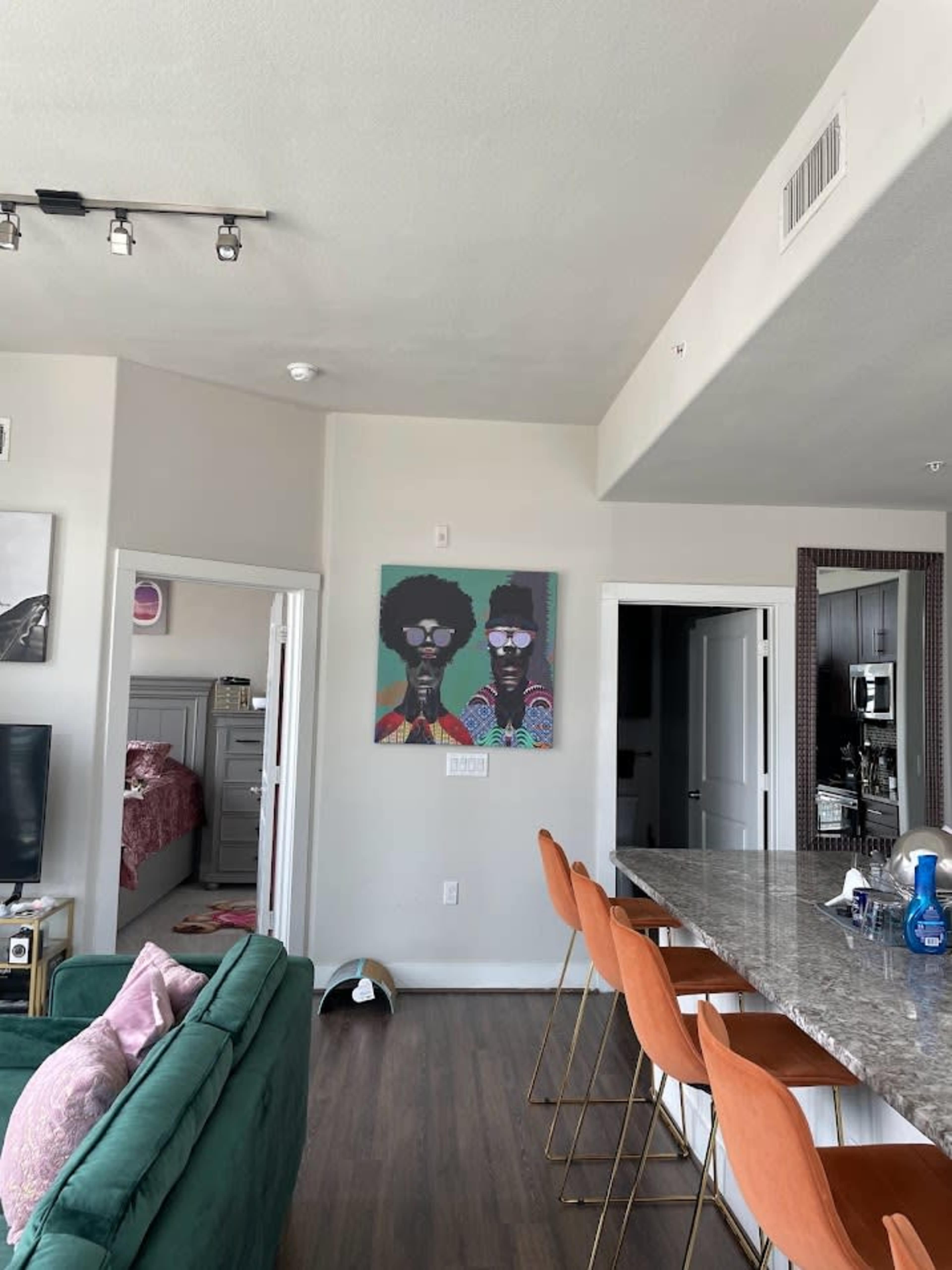 The image shows a modern apartment interior with a green sofa, a kitchen island with orange bar stools, and a colorful artwork of two figures on the wall.