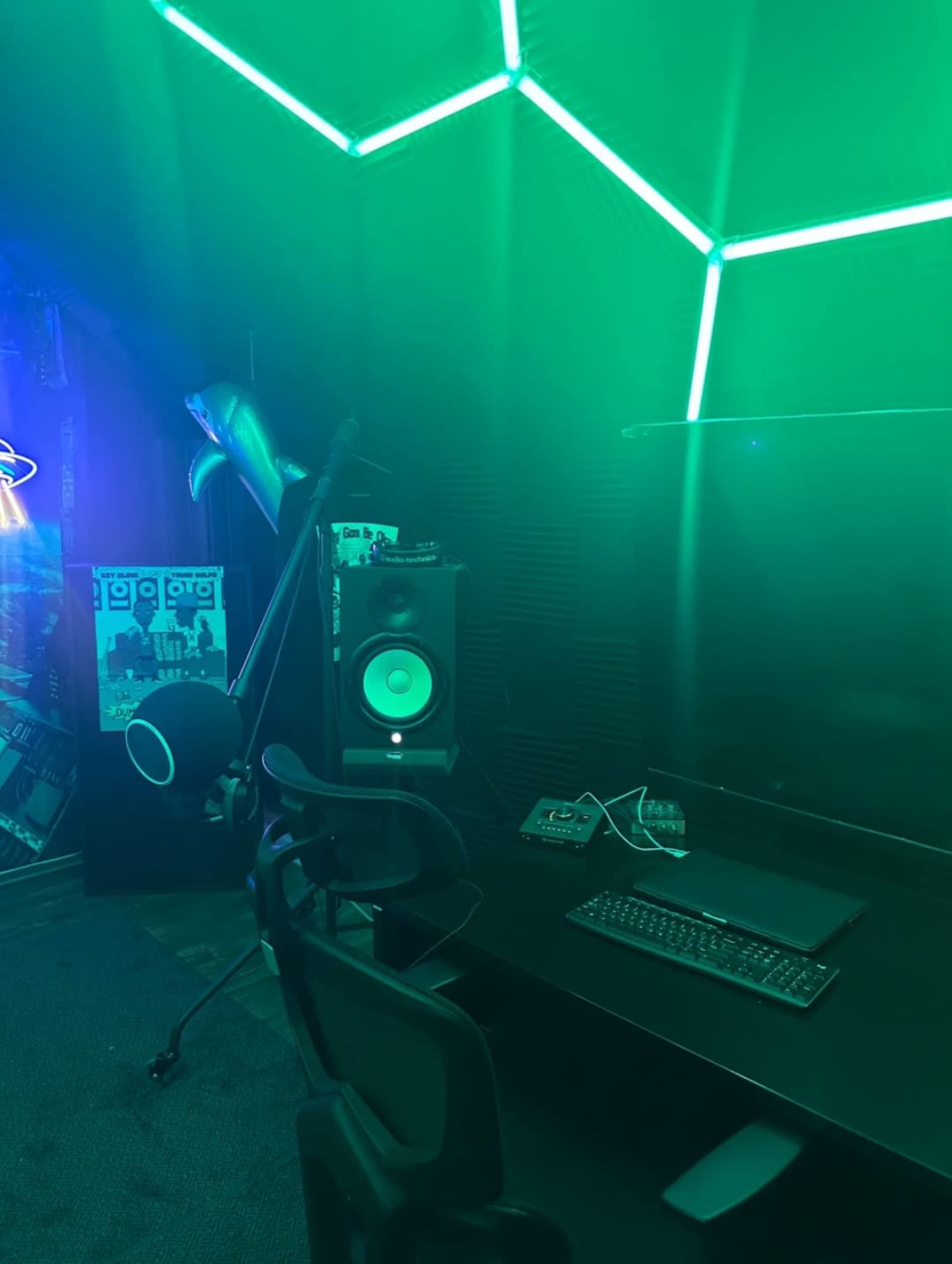 The image shows a dimly lit recording studio with green ambient lighting, featuring a desk with a keyboard and a monitor, a speaker, and a microphone on a stand.
