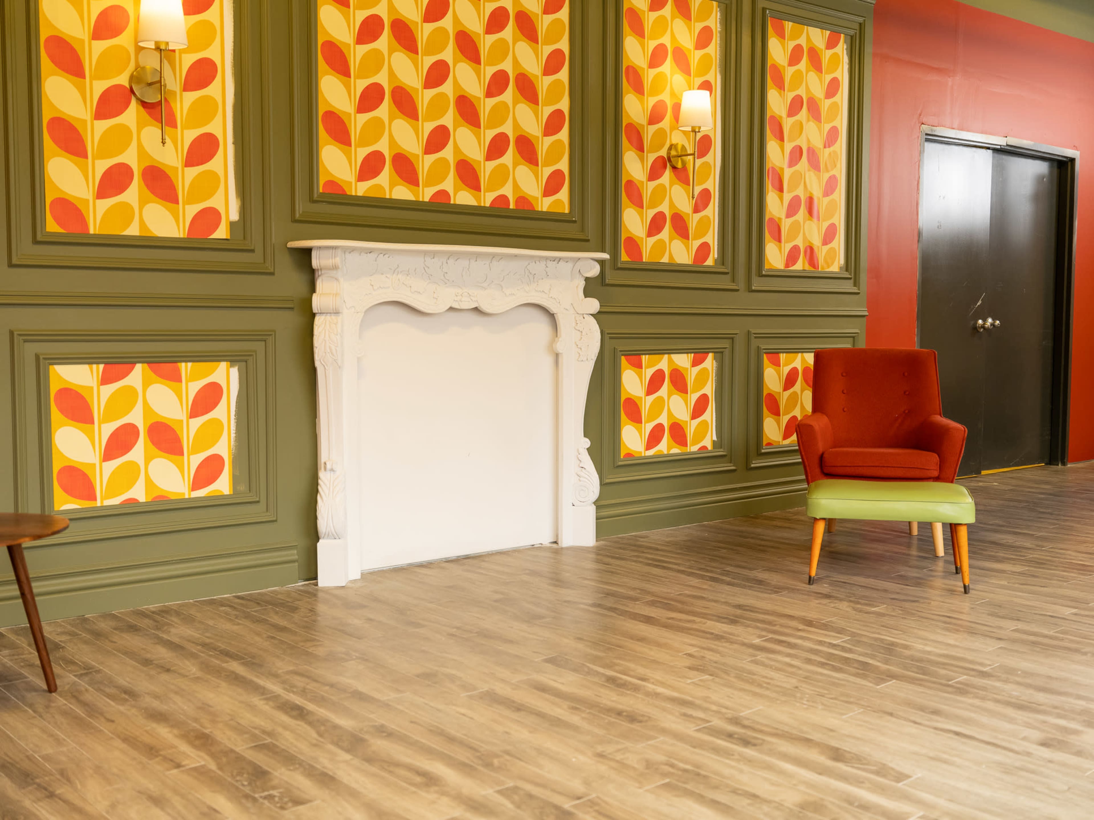 The scene features a room with patterned wallpaper, a white fireplace, and a two-toned armchair against a wooden floor.