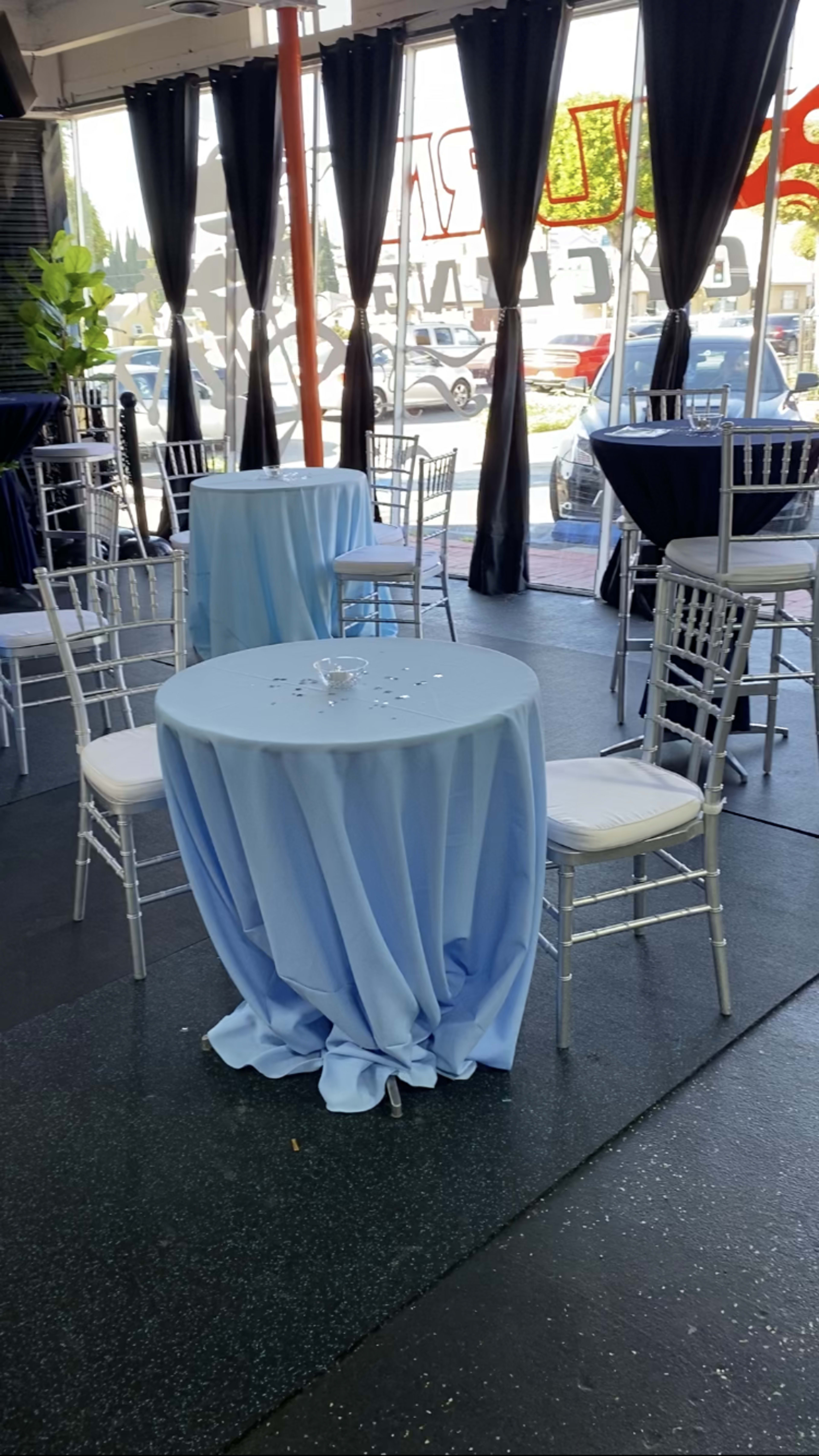 The image shows a small dining area with blue tablecloths draped over round tables, surrounded by silver Chiavari chairs, and large windows displaying the outside view.