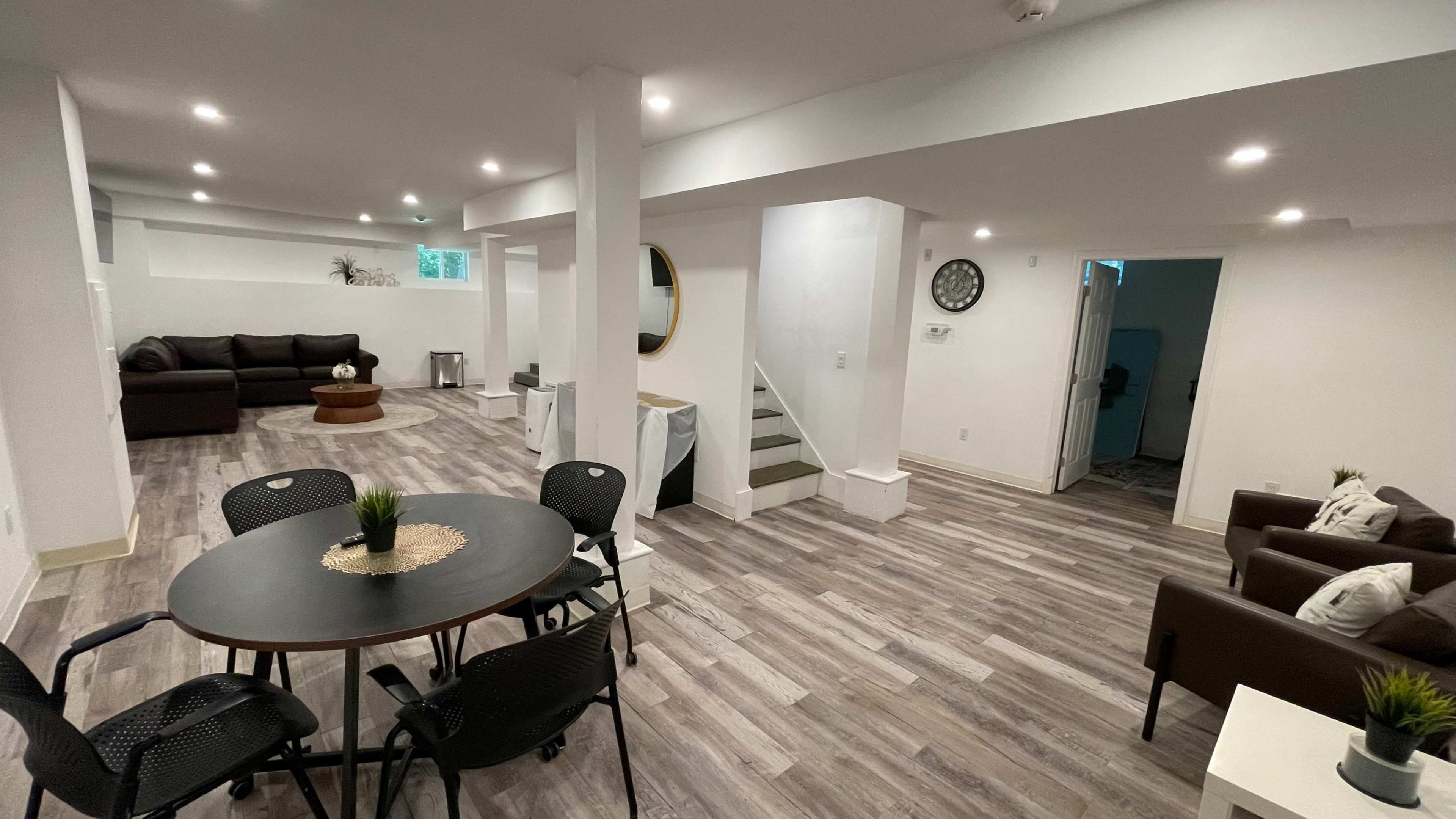 A modern, open-concept basement living space featuring a seating area, a dining table, and light-colored wooden flooring.