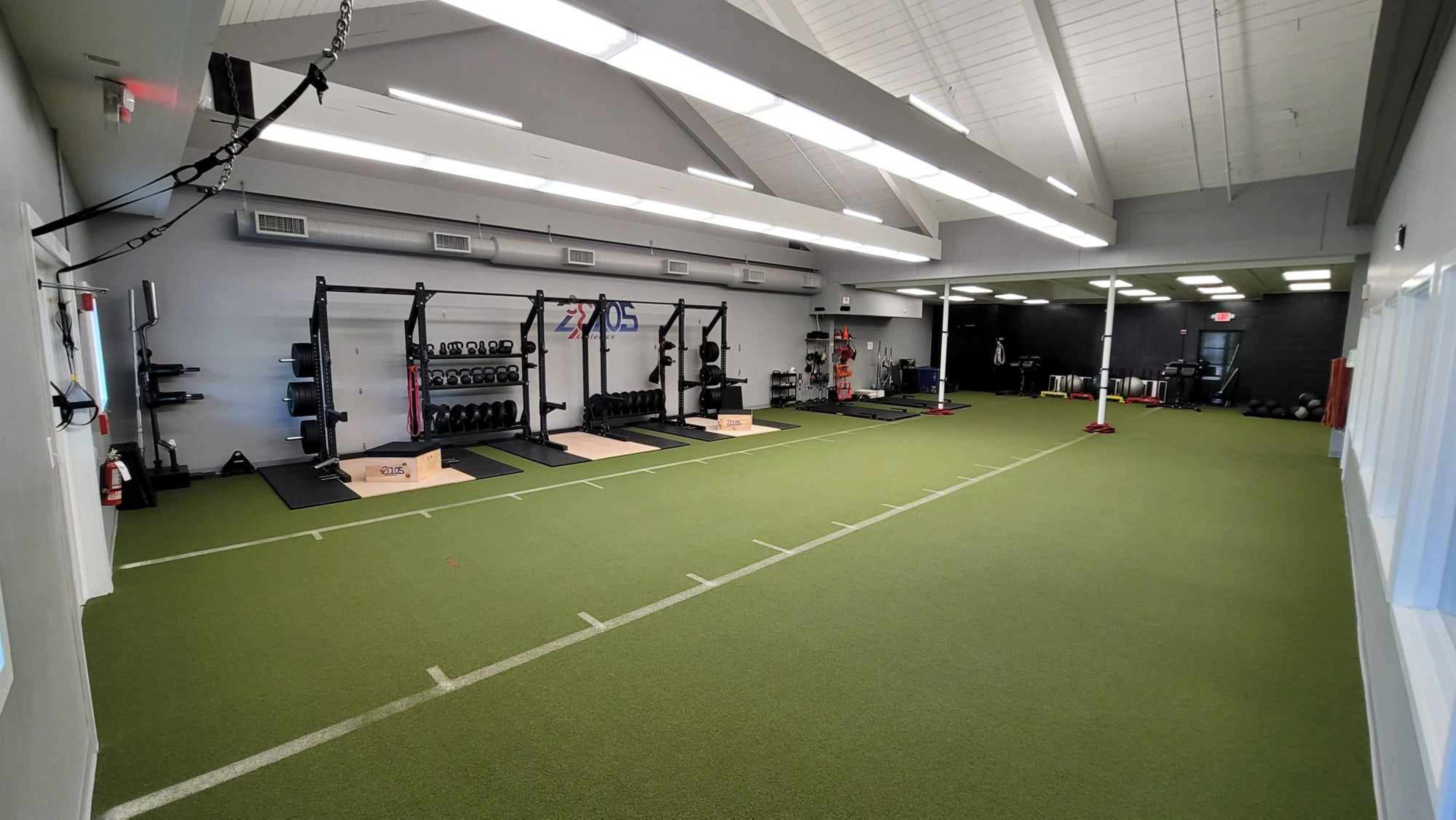 A spacious gym with turf flooring, multiple weightlifting stations, and exercise equipment arranged along the walls.