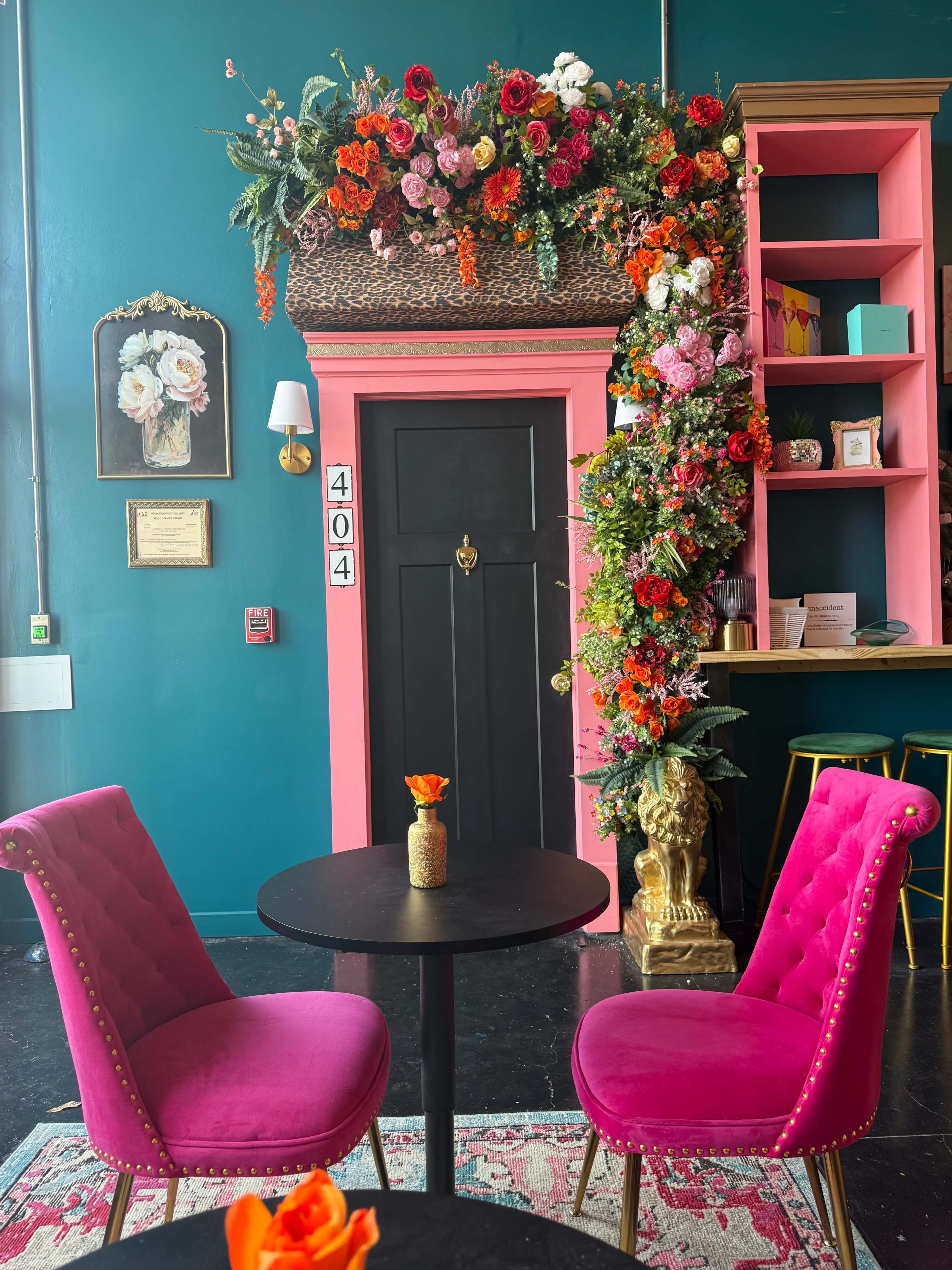 A vibrant café scene features a black door adorned with colorful floral arrangements, flanked by two pink velvet chairs at a small circular table.