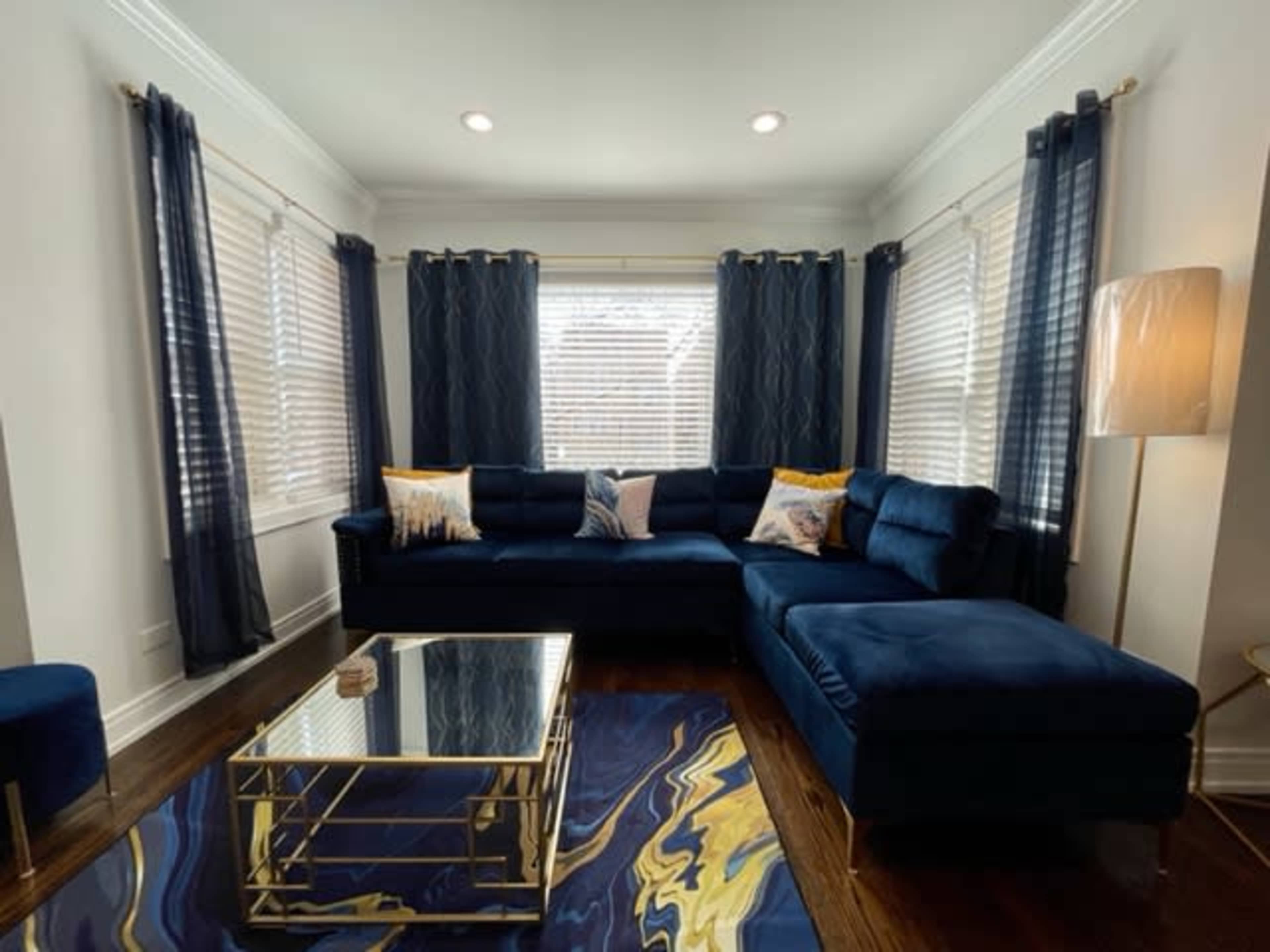The image shows a living room with a blue sectional sofa, gold-accented coffee table, and large windows covered with dark curtains.