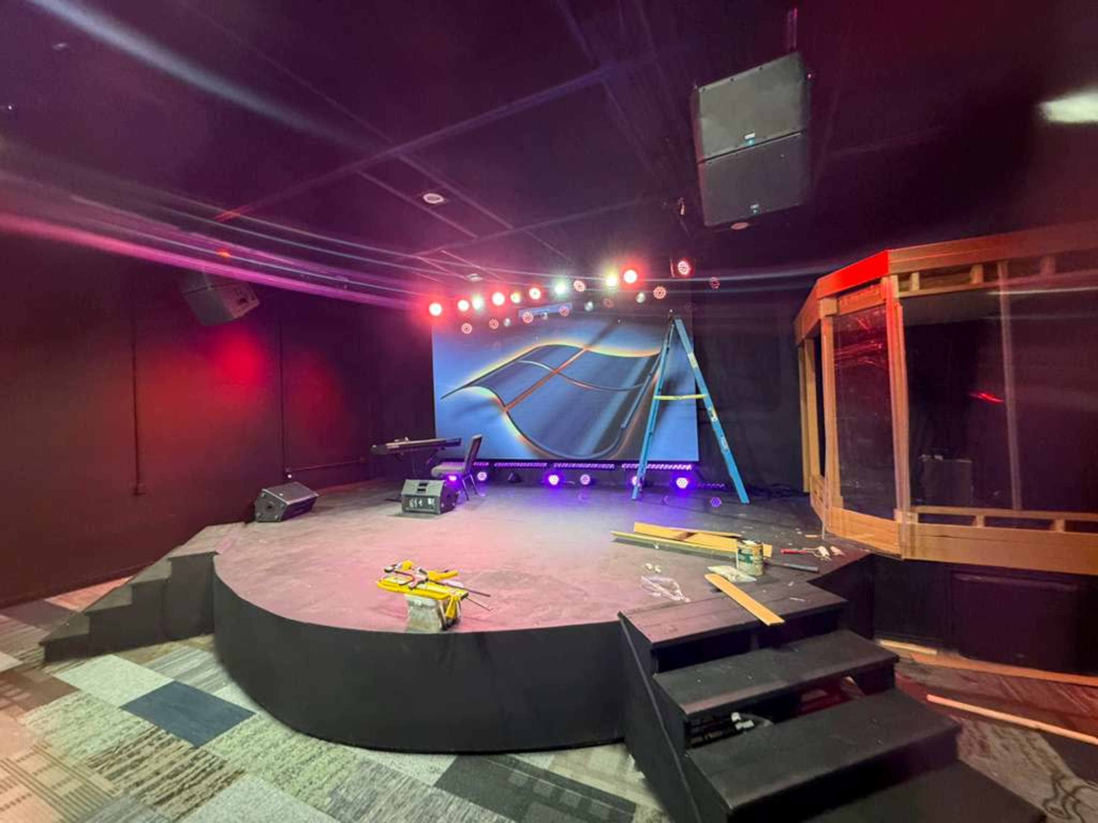 A partially set-up stage features a curved platform, a backdrop with abstract designs, and various lighting fixtures in place, alongside construction materials scattered on the floor.