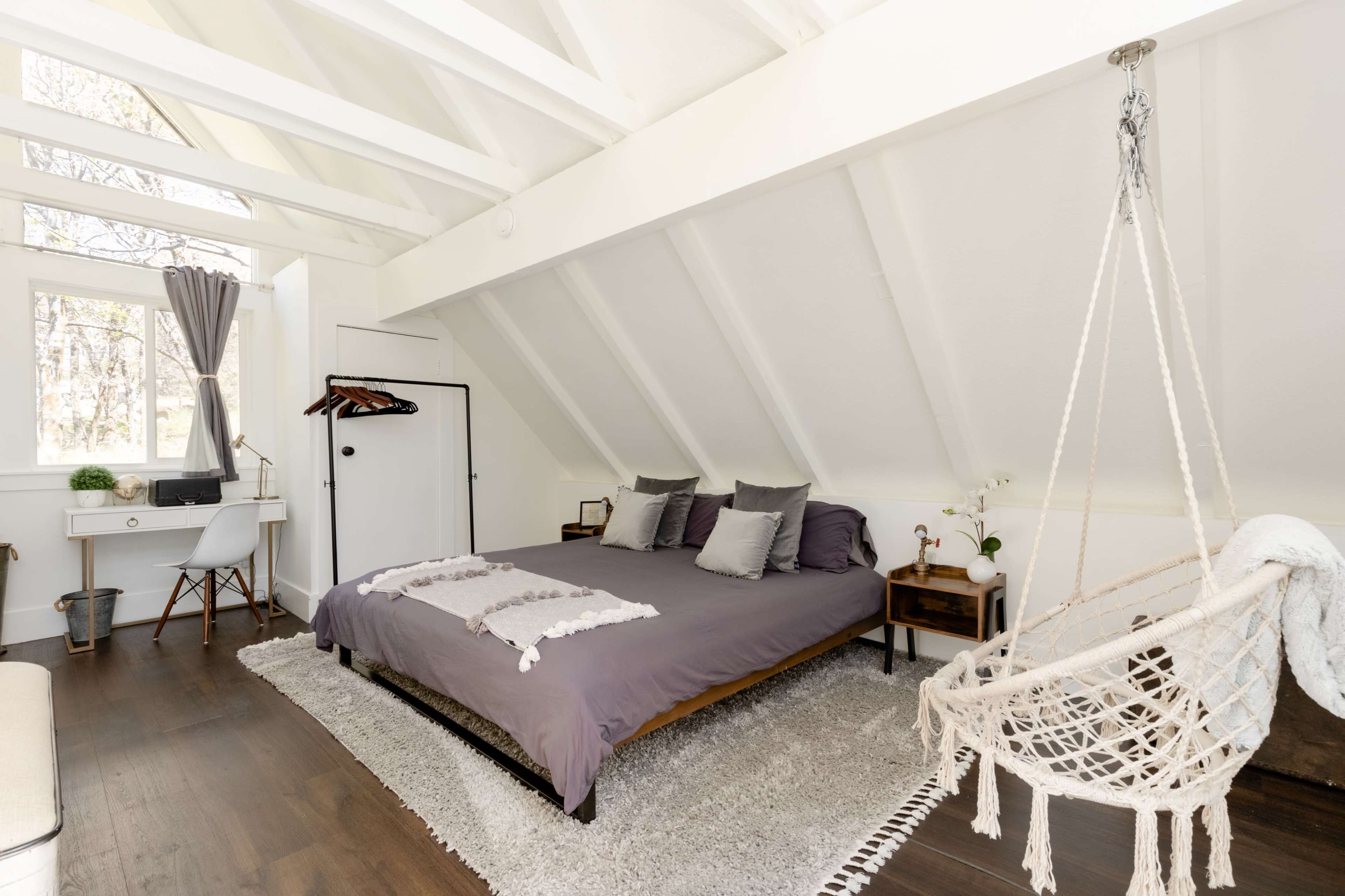 The image shows a minimalist bedroom with a large bed, a desk and chair, and a hanging macramé swing.