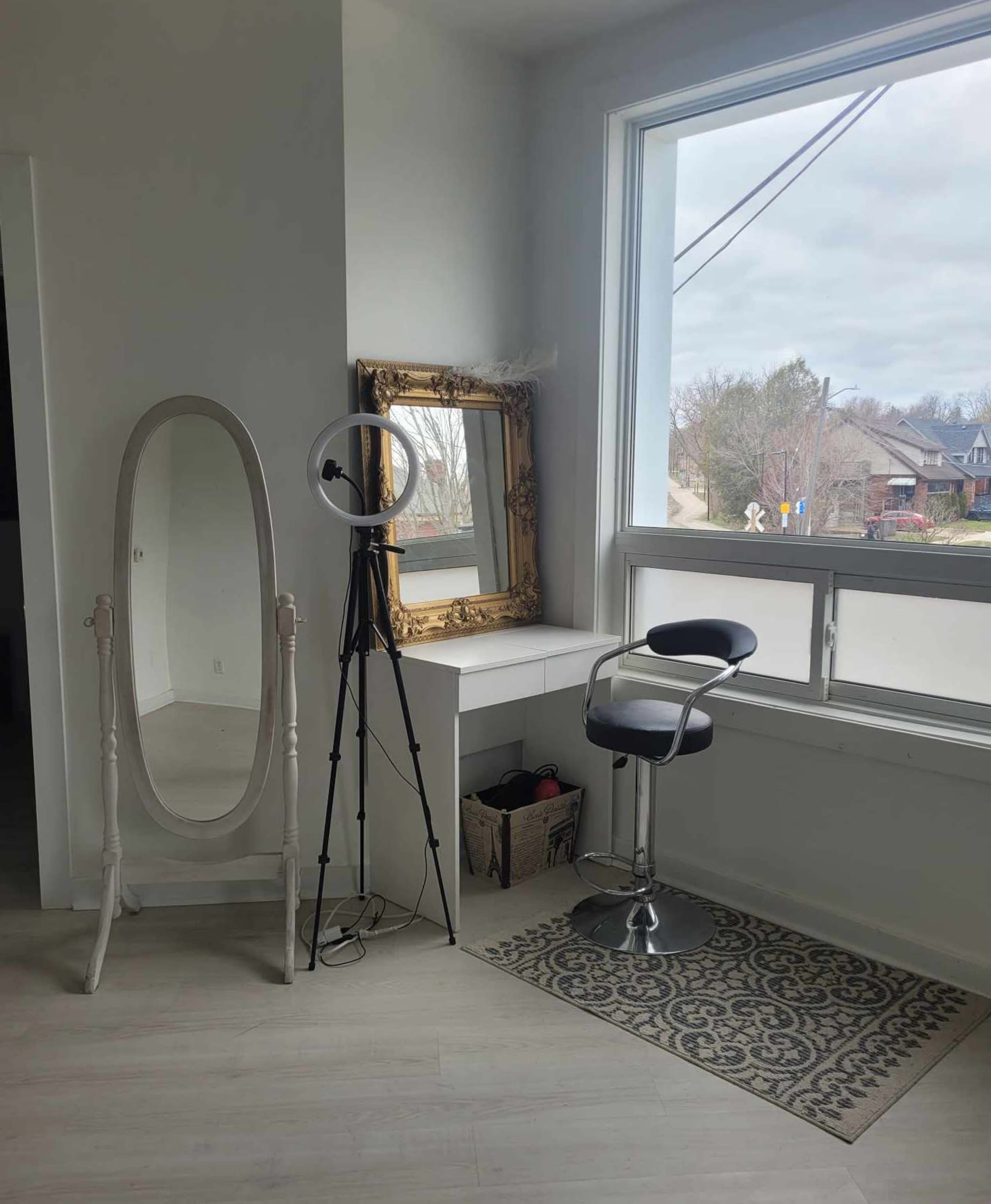 A bright room featuring a standing mirror, a ring light on a tripod, a modern bar stool, and a small desk positioned near a large window.