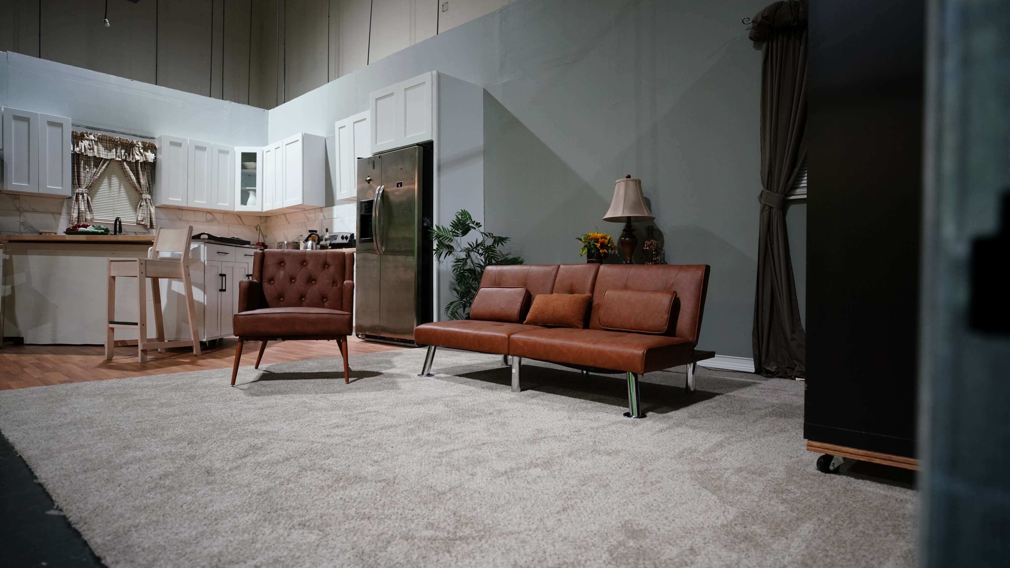 The image shows a modern living room setup with a brown leather sofa and a matching armchair on a light-colored rug, adjacent to a kitchen area with white cabinetry and appliances.