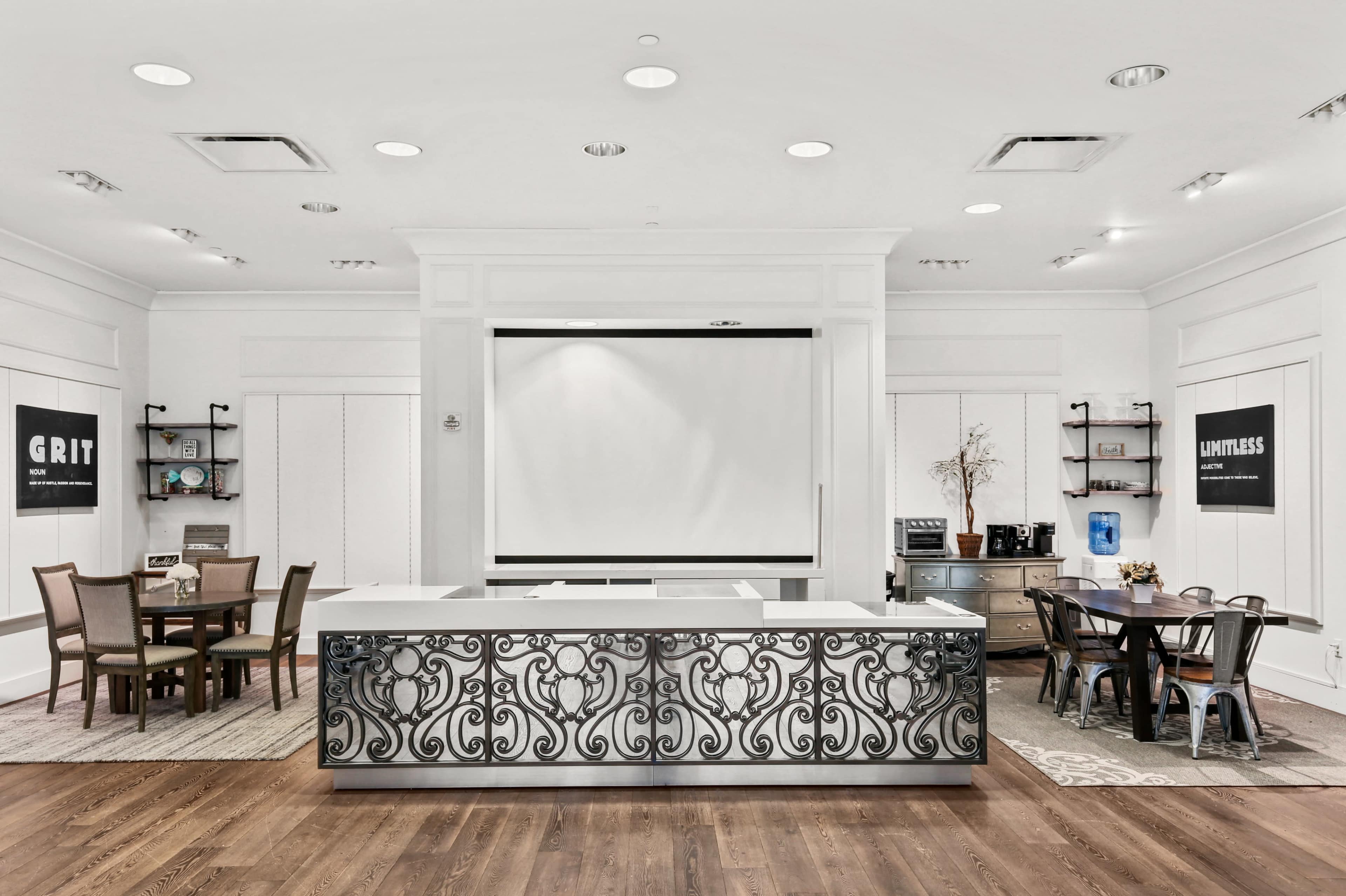 A modern kitchen and dining area with a central island featuring ornate metalwork, surrounded by tables and chairs against a backdrop of white walls and framed signs.