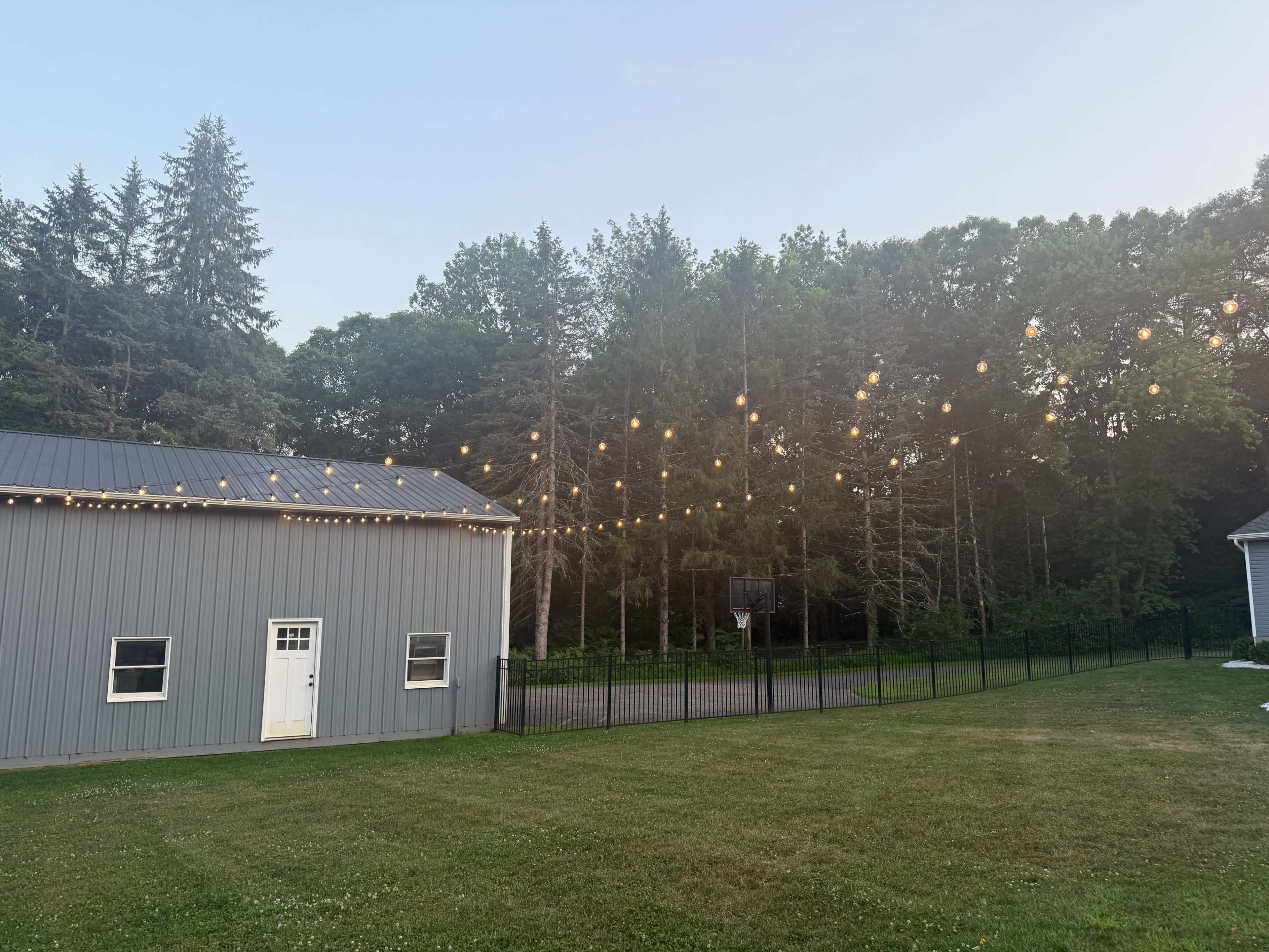 A gray metal building is situated next to a grassy area, with string lights overhead and a basketball hoop visible in the background near a tree line.