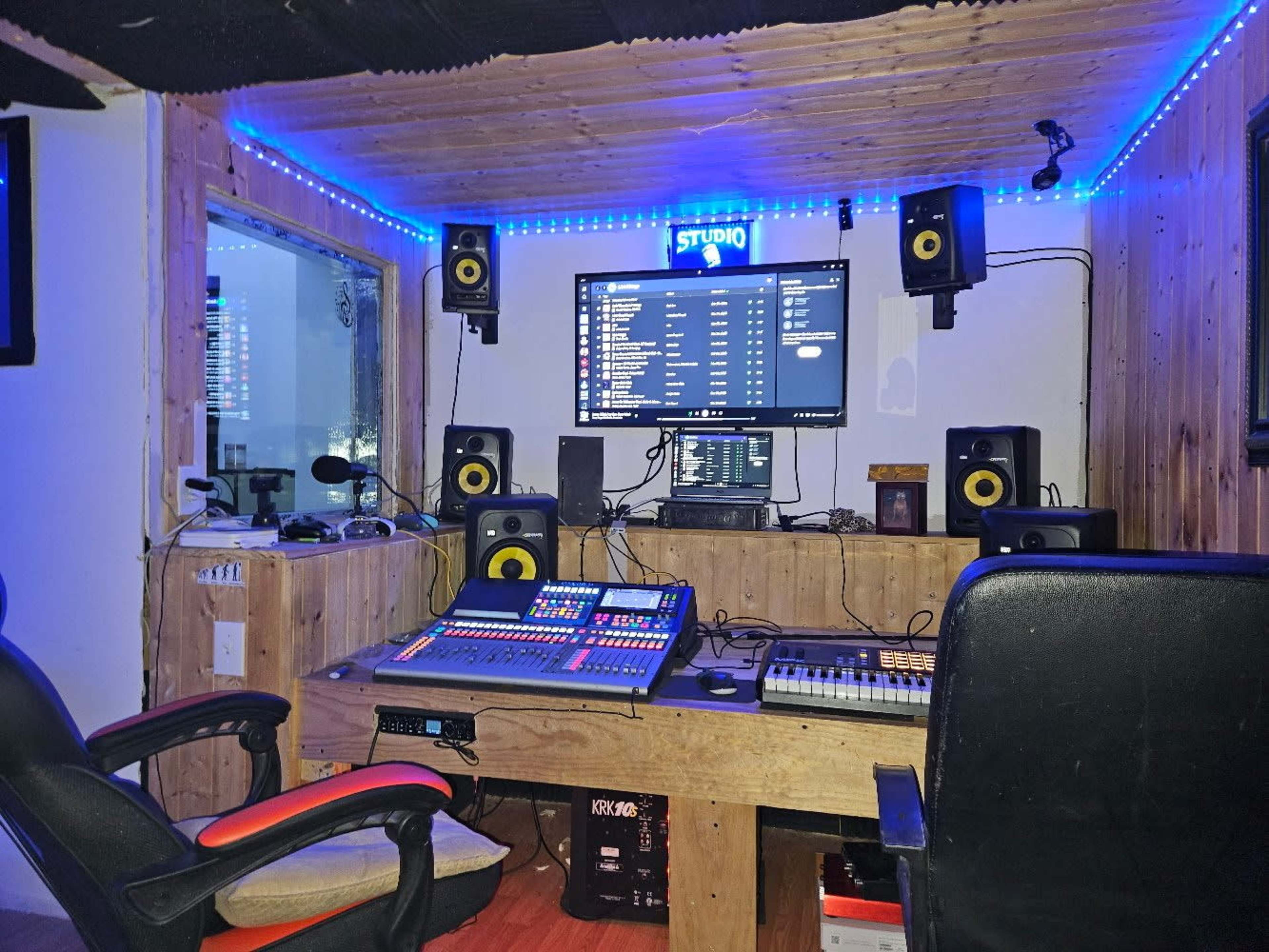 The image shows a recording studio with a wooden interior, featuring a large mixing console, computer monitor, and sound monitors mounted on the walls, illuminated by blue LED lights.