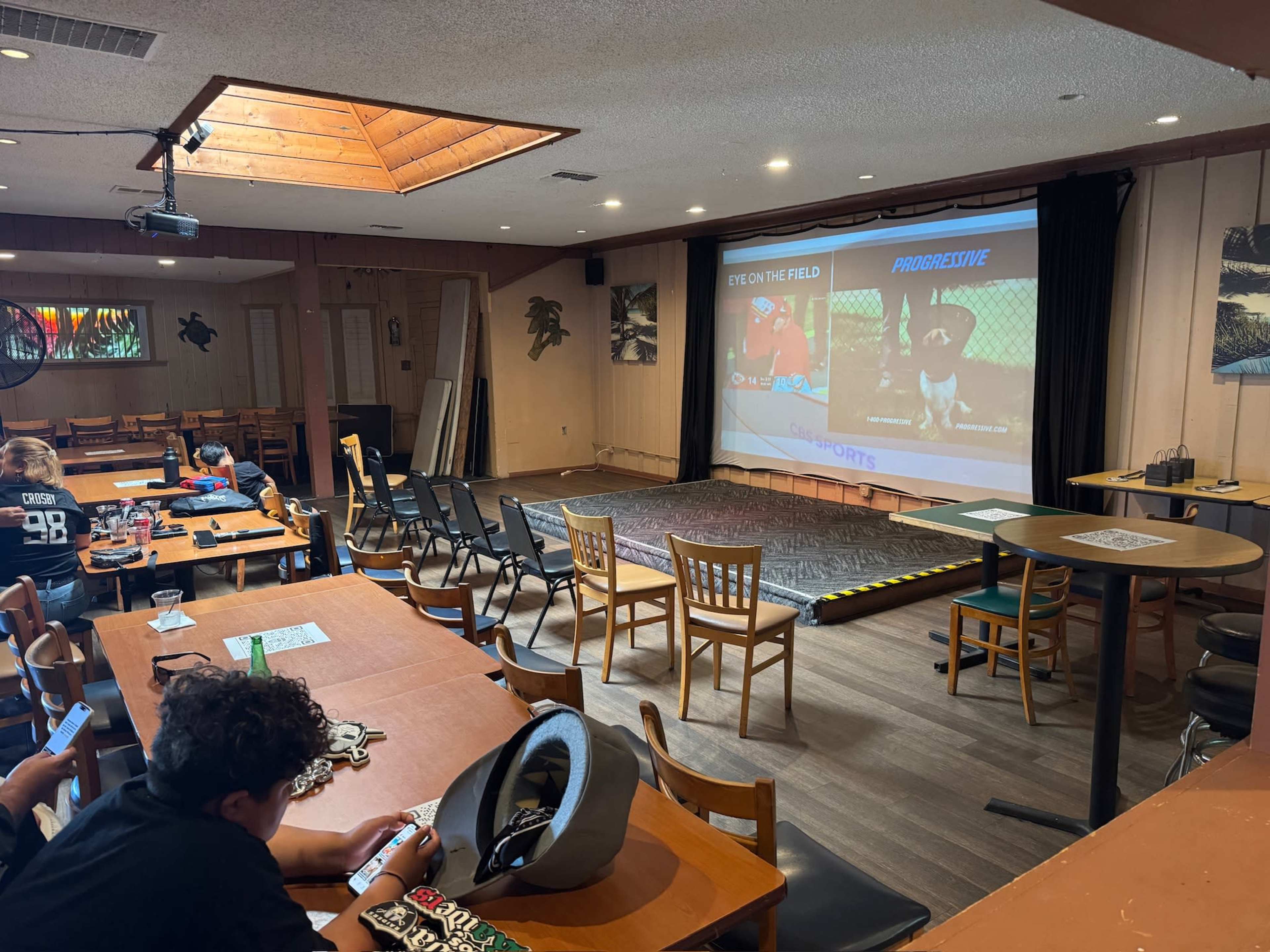 The image shows an indoor sports-themed venue with tables, chairs, a large screen displaying a sports video, and several people seated, some using their phones.