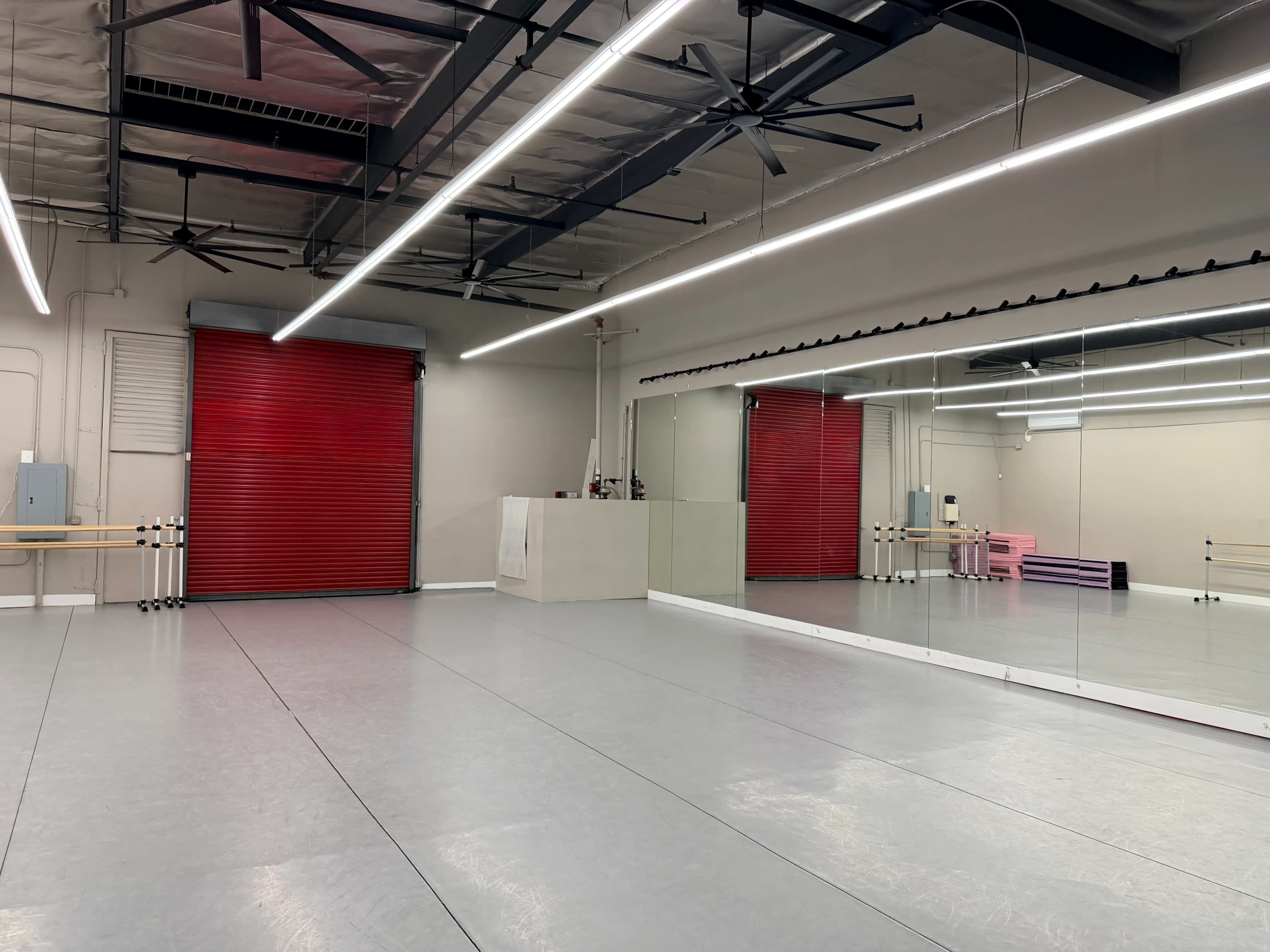 The image shows a spacious dance studio with gray flooring, large mirrors on one wall, and a red roll-up door at the opposite end.