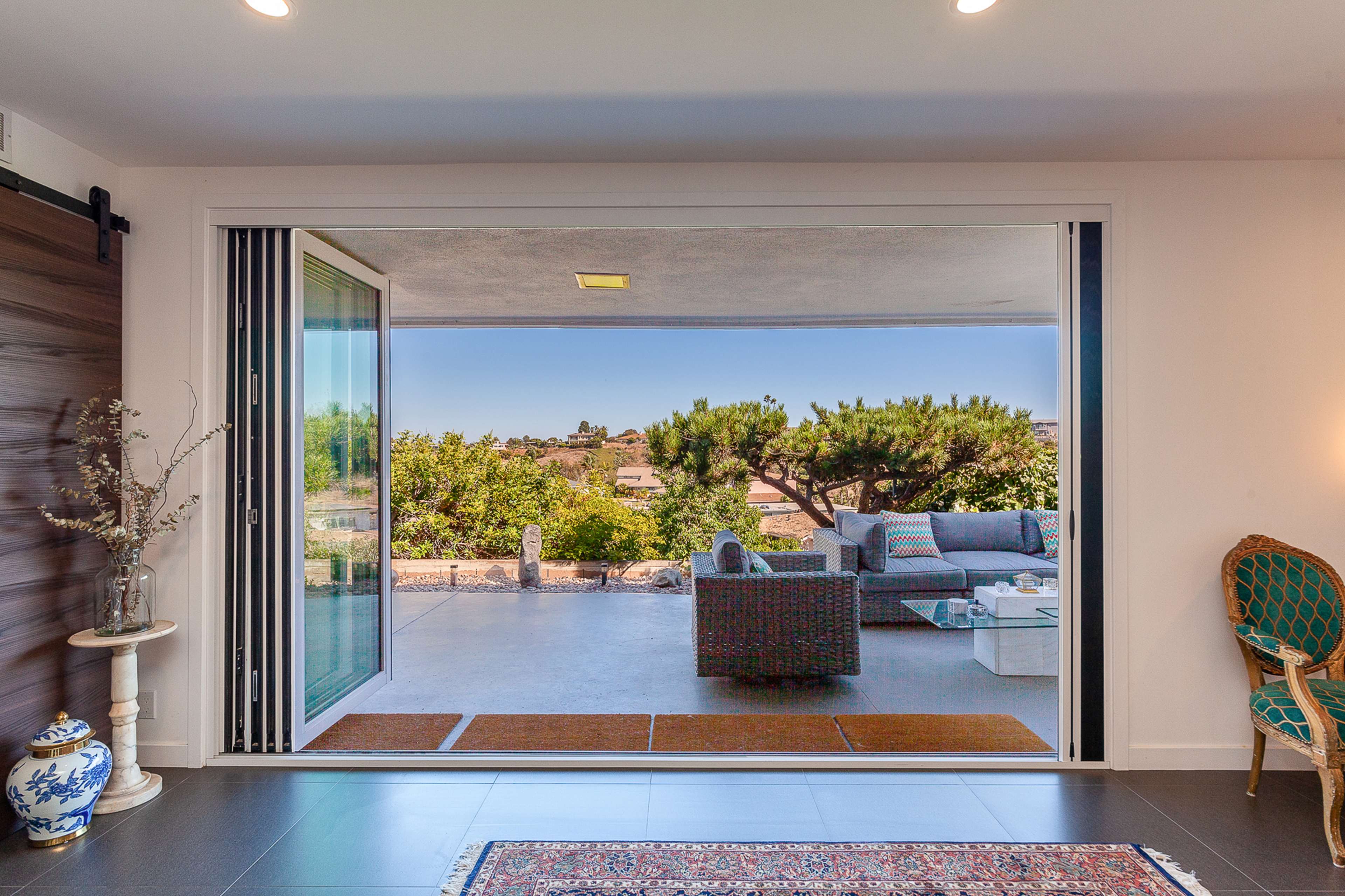 A spacious living area opens to a patio with outdoor seating and a view of greenery and distant hills.