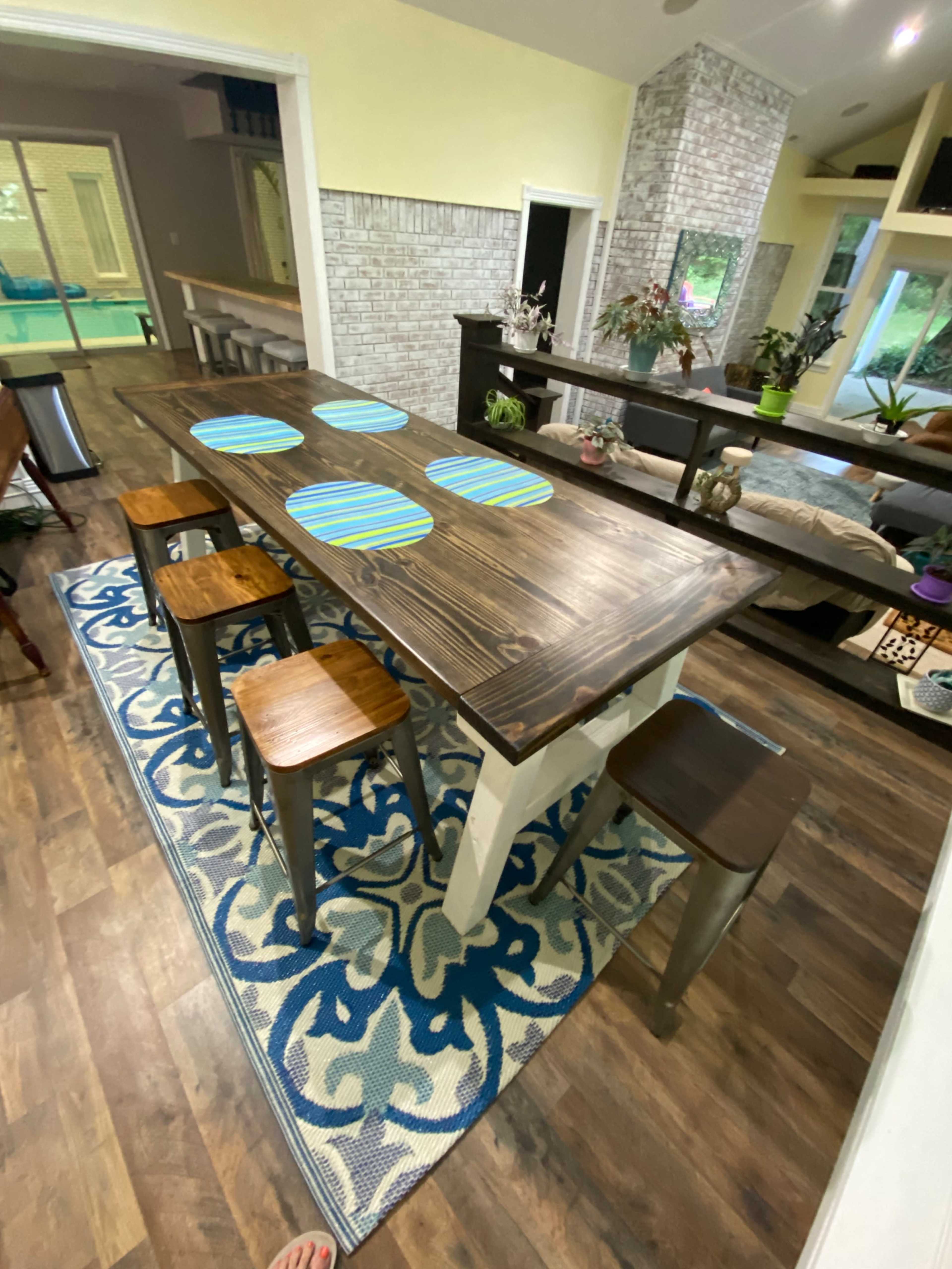 A large wooden dining table with striped placemats is surrounded by four stools in a brightly lit room with a patterned rug.