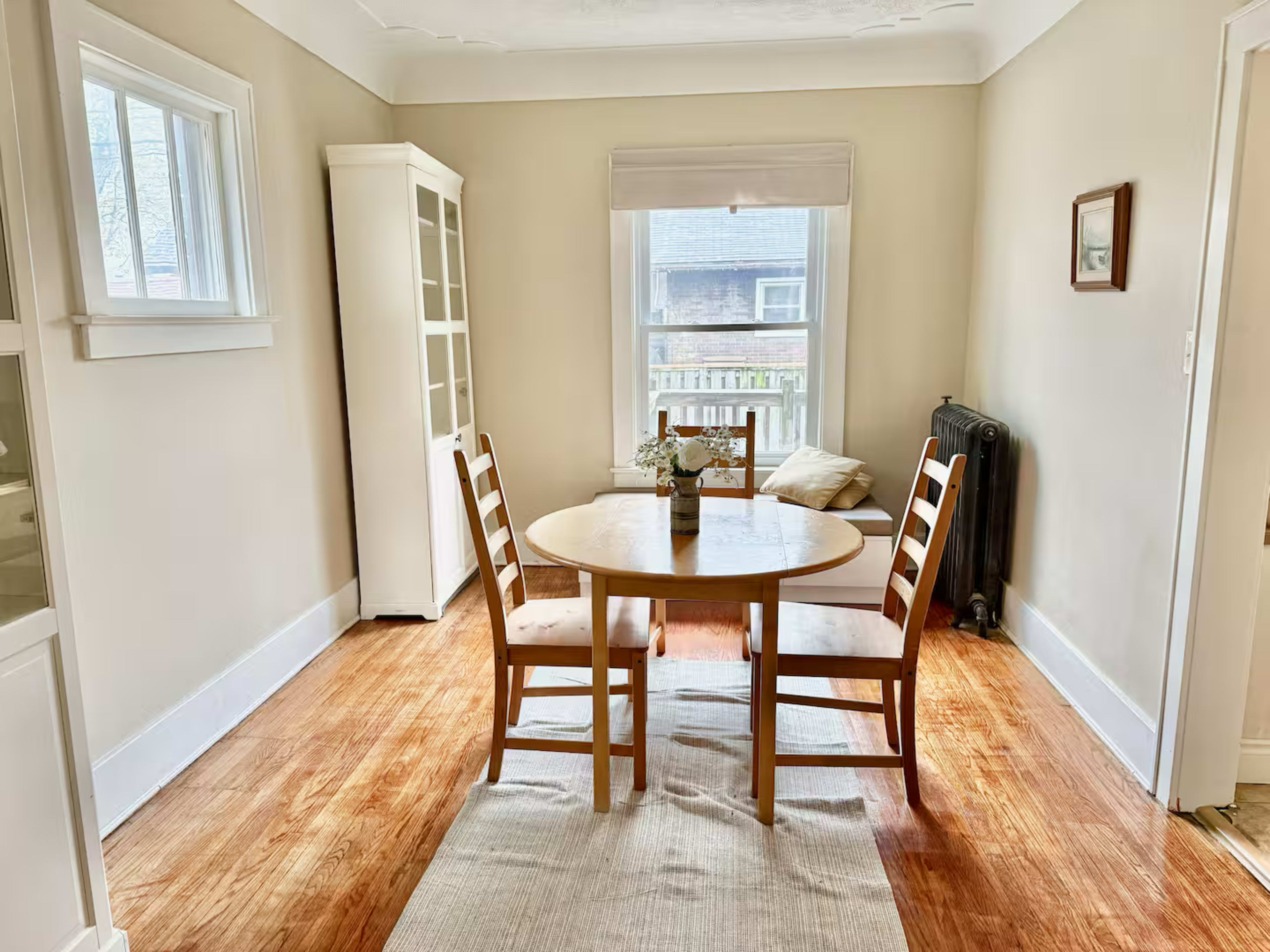 A small dining area features a round wooden table with two chairs, a vase of flowers, and a window overlooking an exterior view.