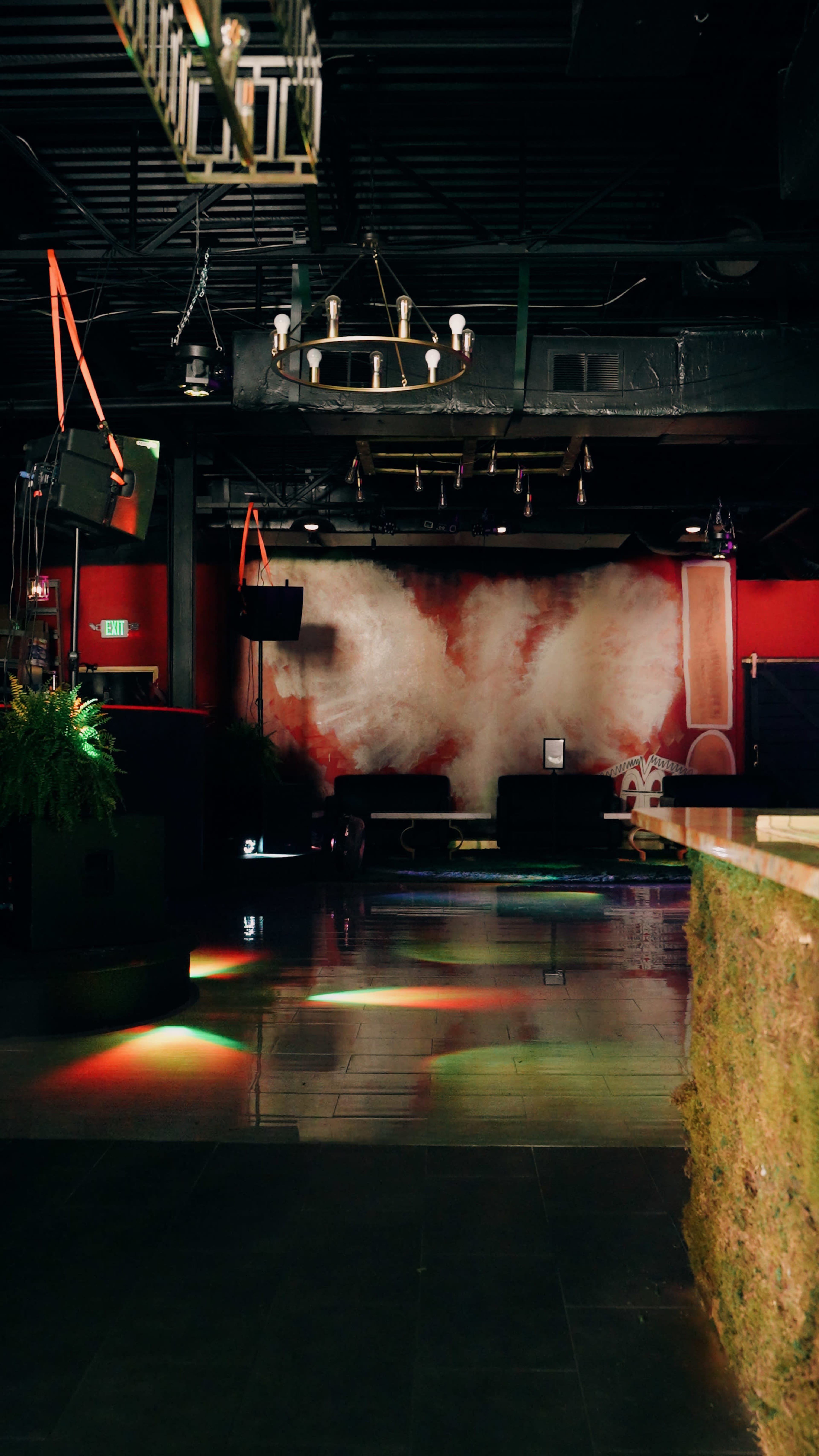 The image shows an empty indoor venue with a colorful mural on the wall, dim lighting, and a bar area featuring a moss-covered surface.