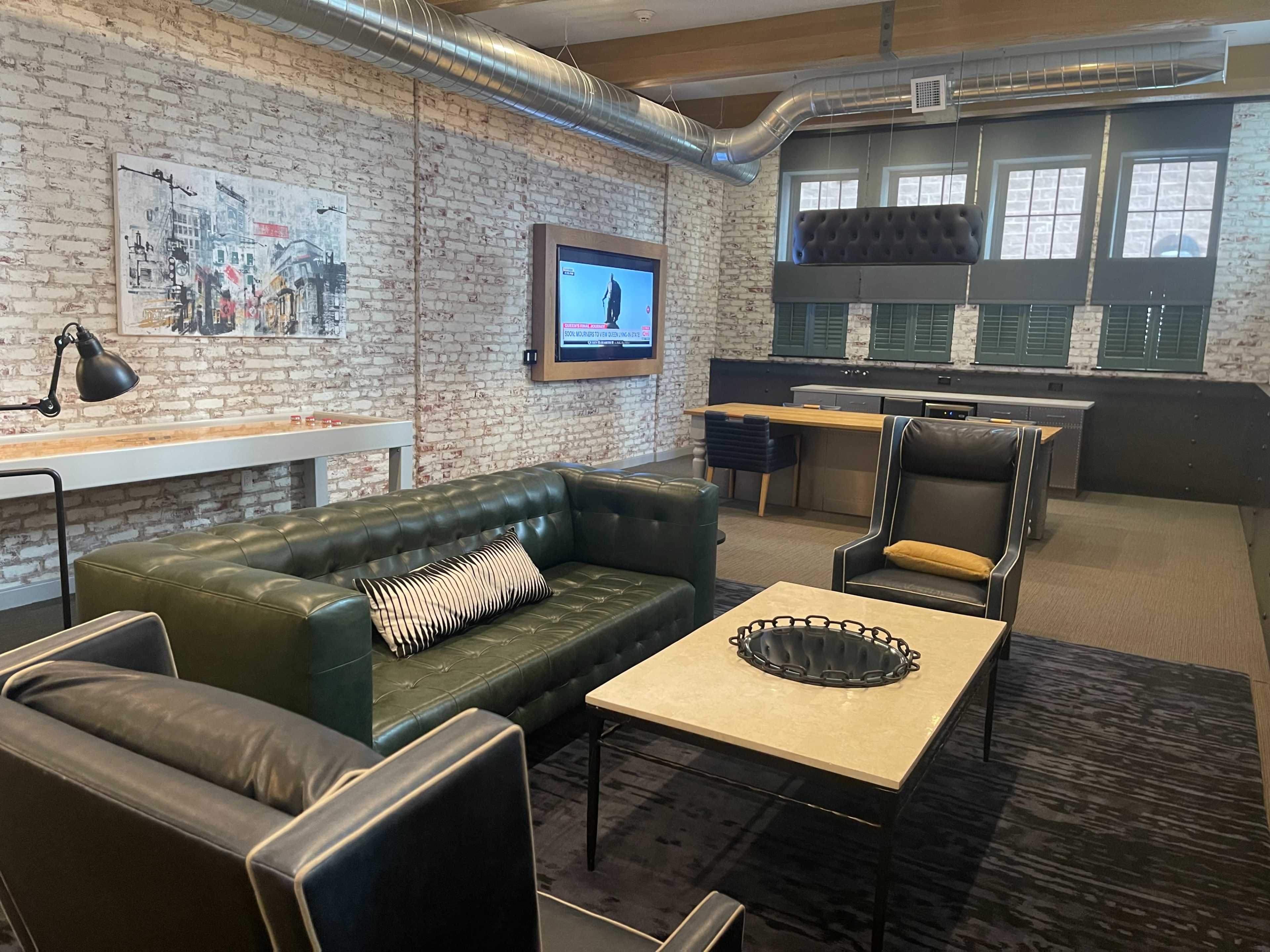 A modern lounge area with brick walls, two green sofas, a coffee table, a wall-mounted television, and a bar counter at the back.