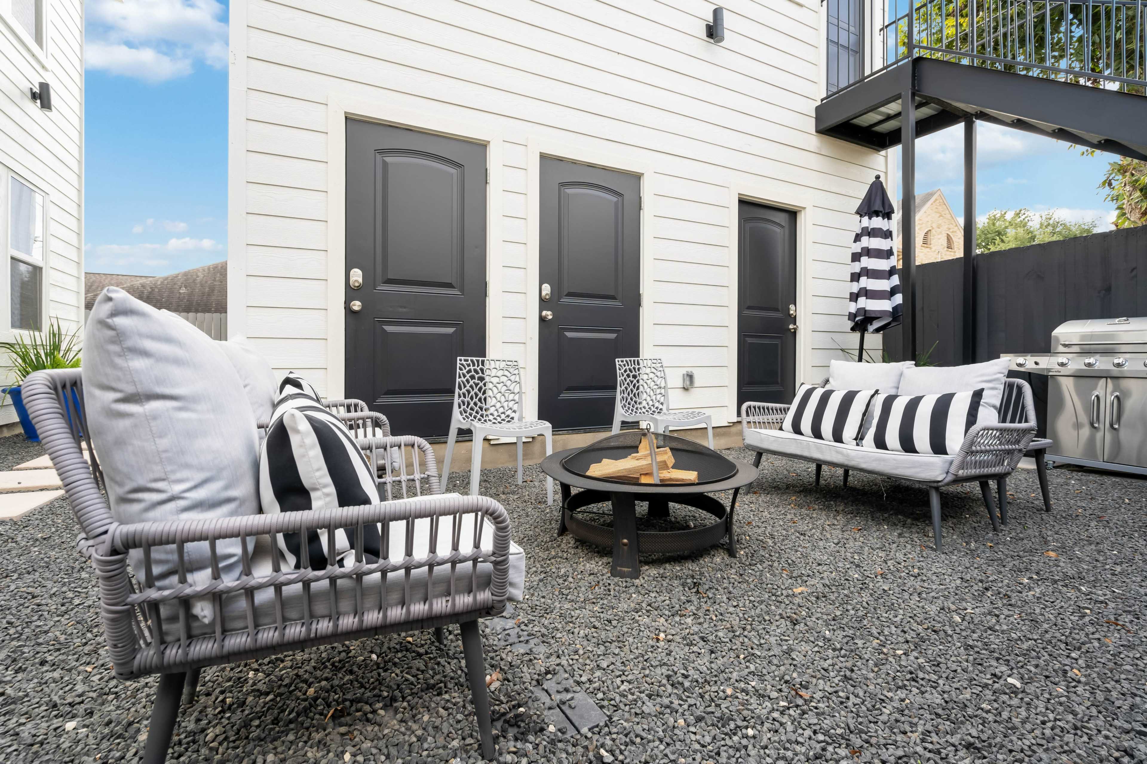 A small outdoor seating area with two couches, a round coffee table, and an umbrella, surrounded by gravel and adjacent to a building with three doors.