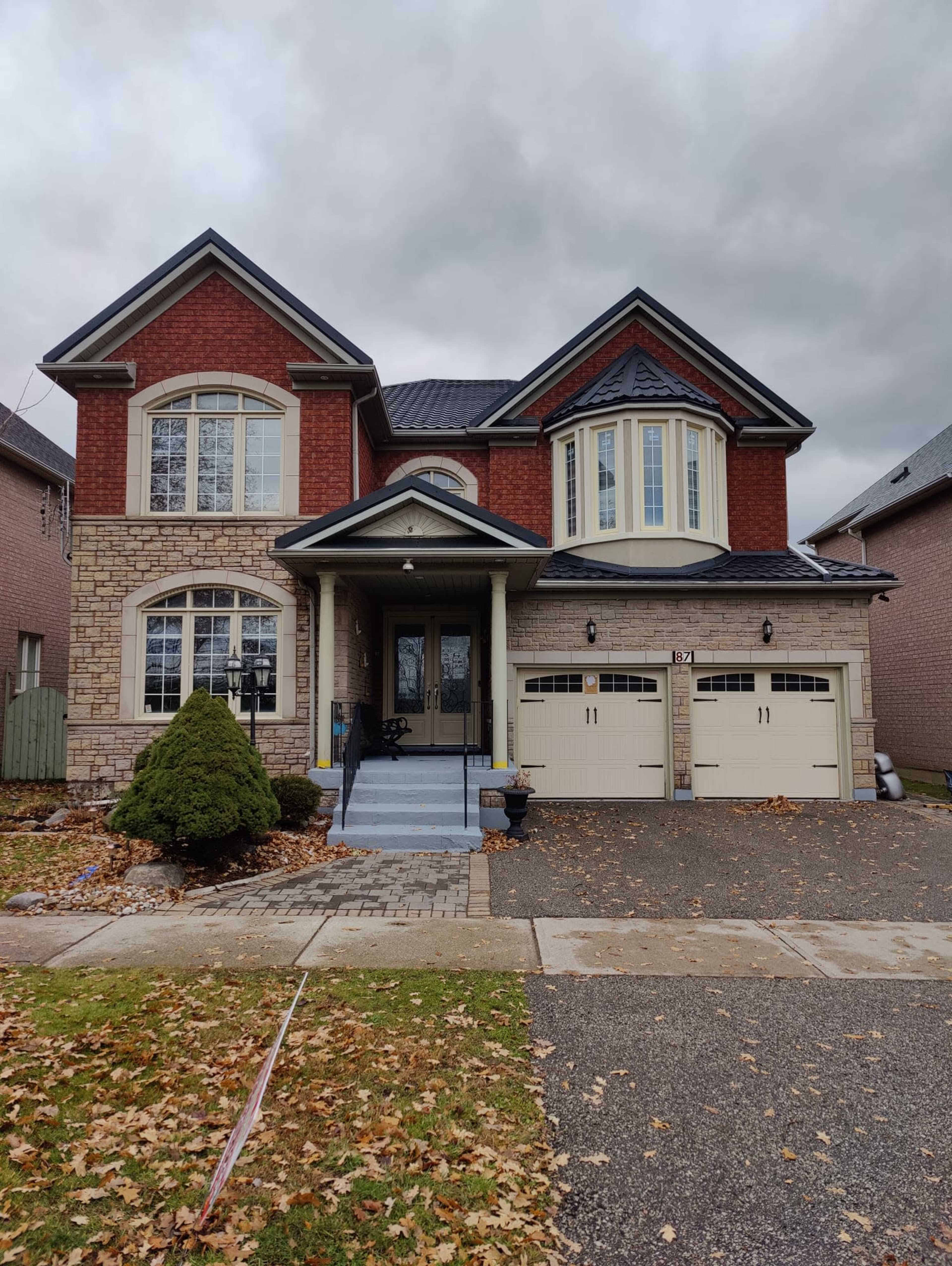 A two-story brick house with a front porch and a double garage sits on a leaf-strewn driveway.
