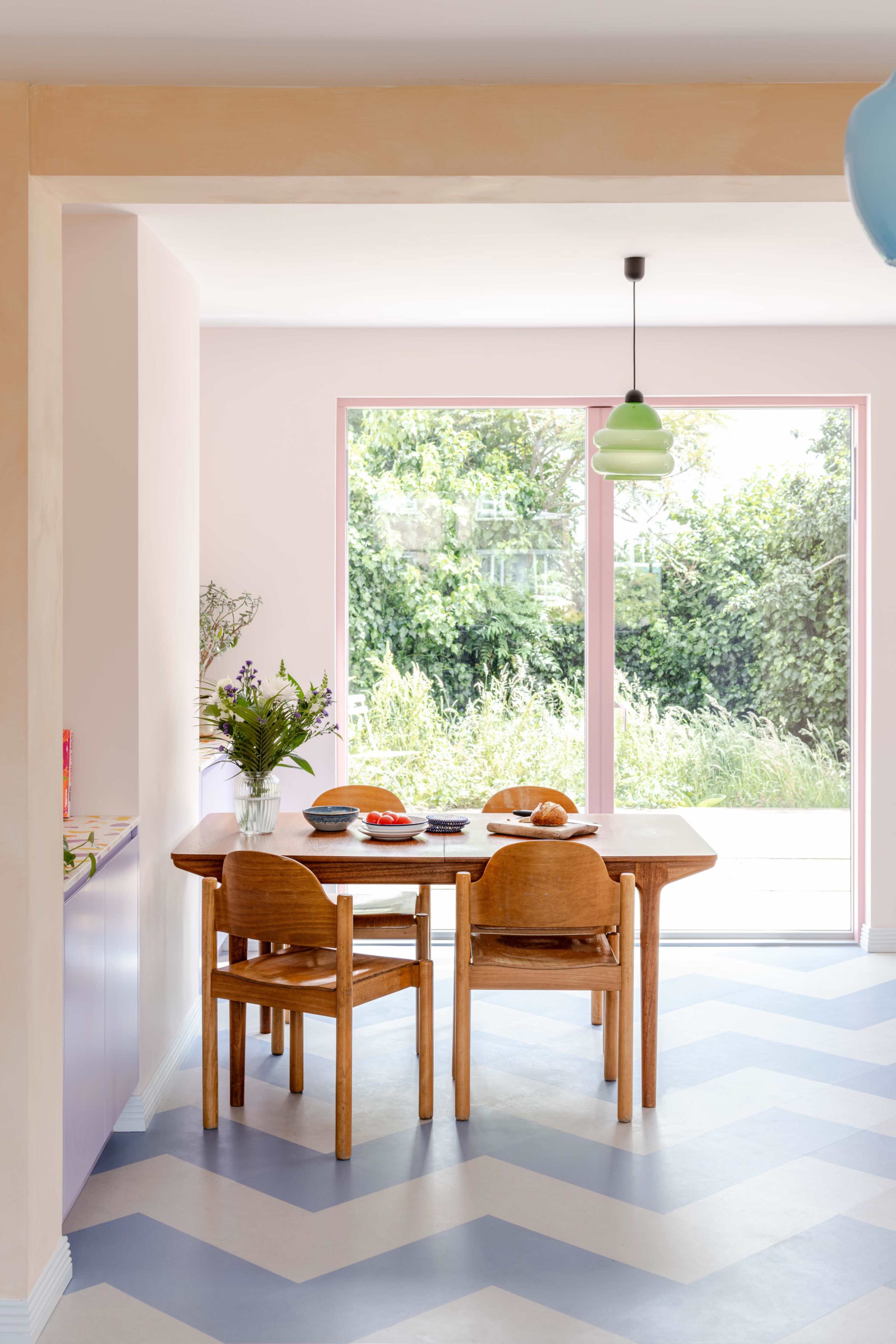 A wooden dining table with chairs is set in a light pink room, adjacent to a large window overlooking greenery outside.